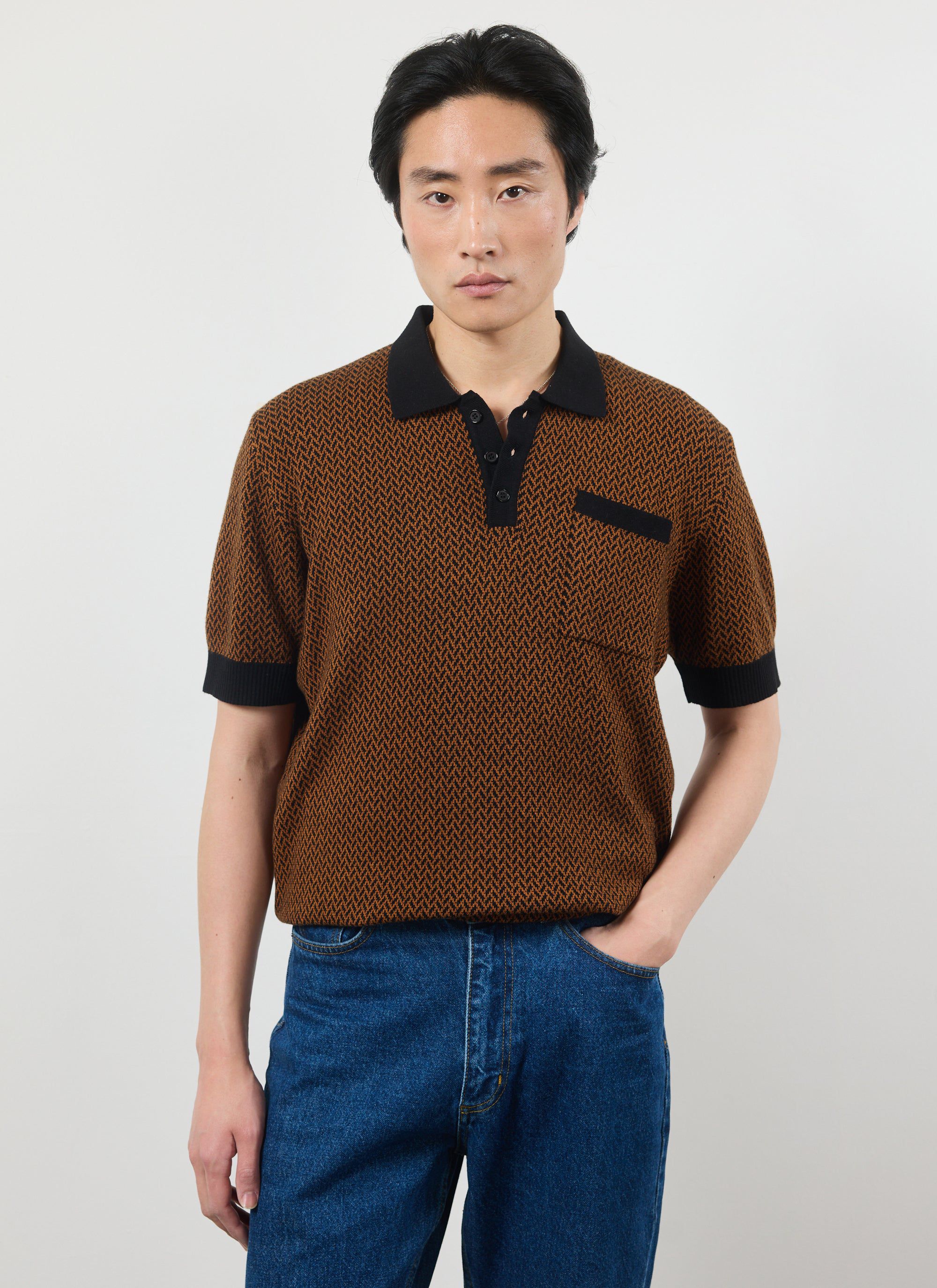 A model wears the Percival Casa Martini Knitted Polo in Espresso, a brown and black patterned short-sleeve shirt with jeans.
