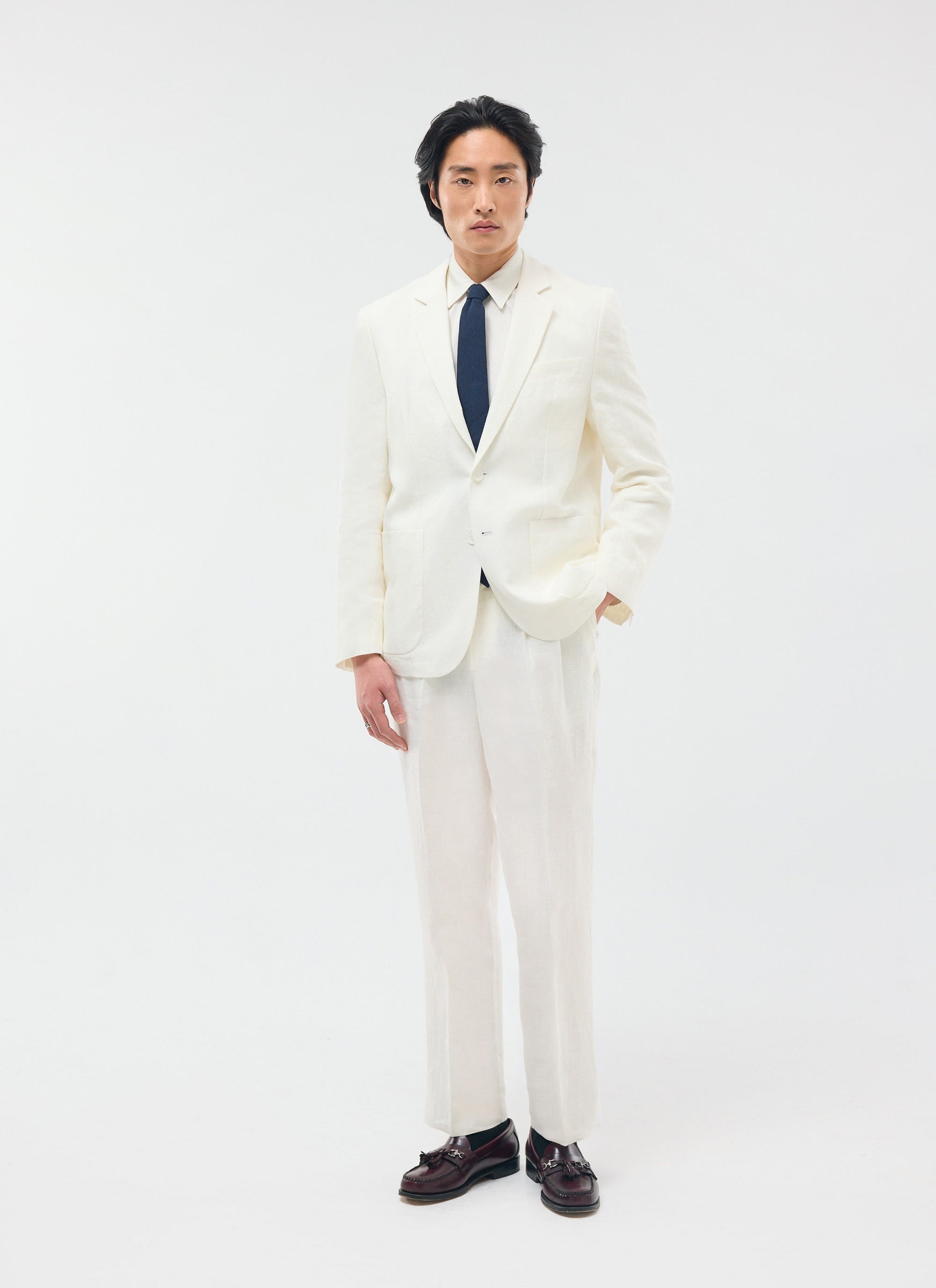 A model wears the full Percival Relaxed Tailored Linen Suit in white, styled with a navy tie and brown loafers.