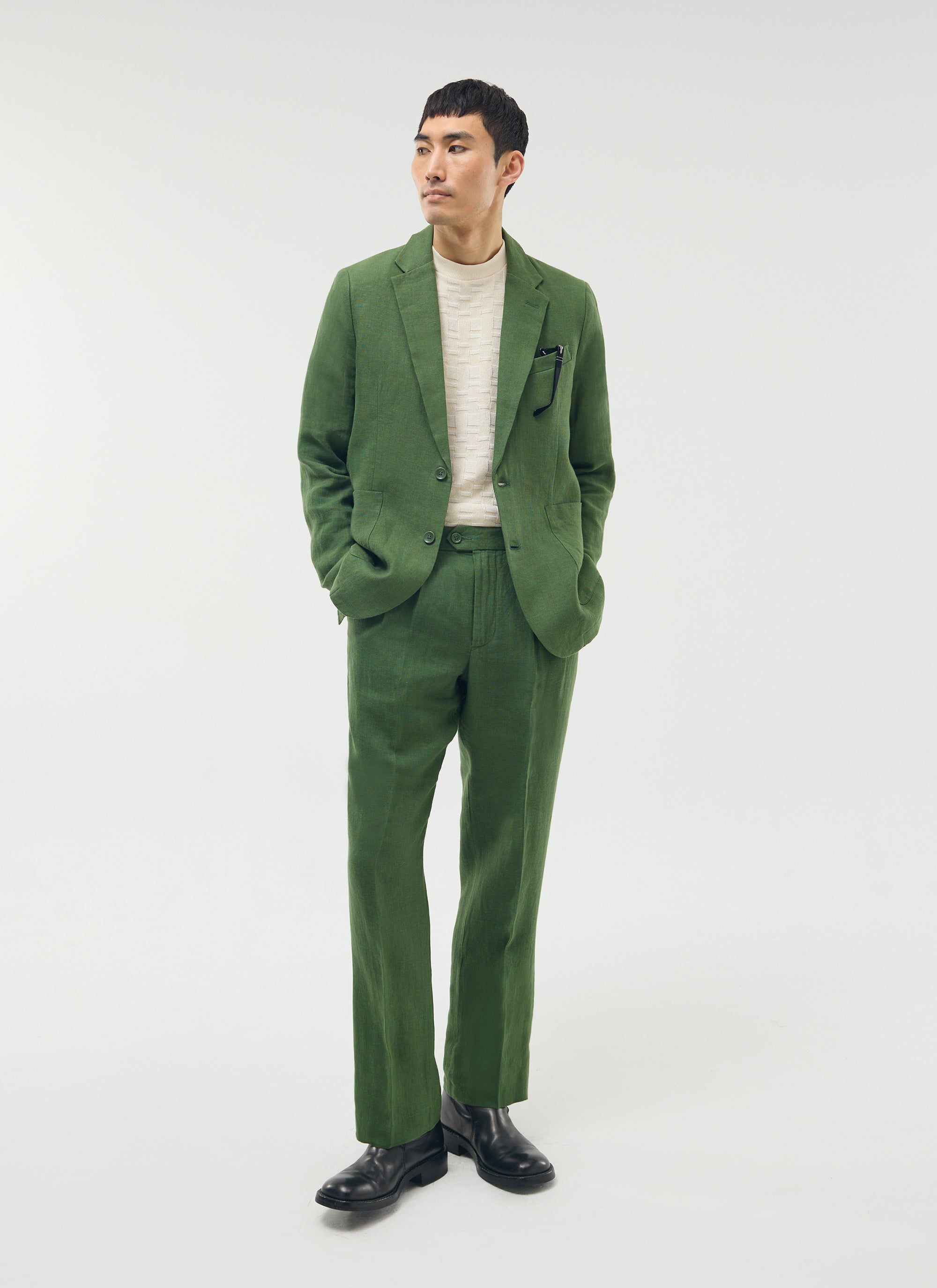 A model wears Percival's forest green linen pleated trousers with the matching blazer, a cream knit and black boots.