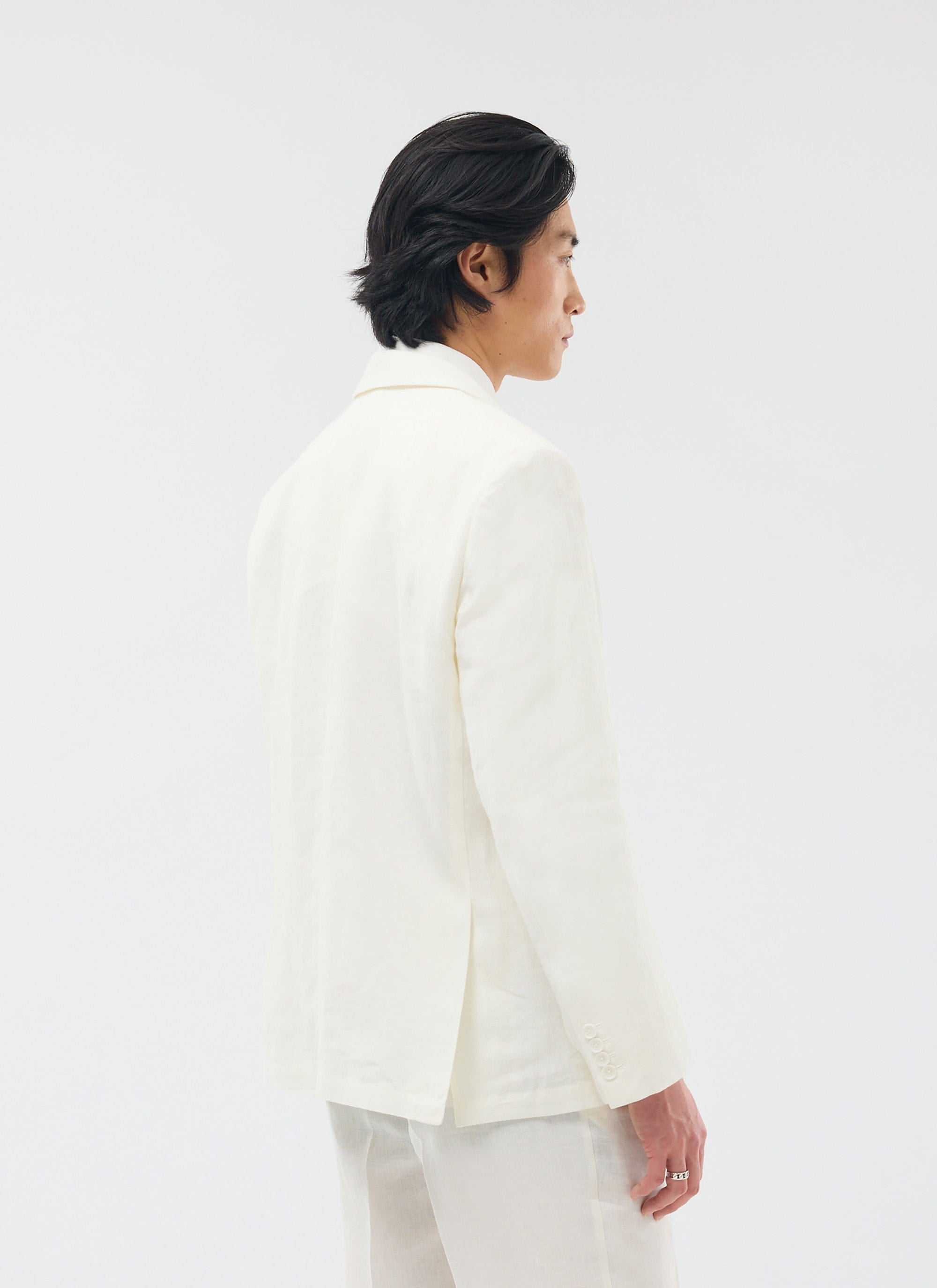 A model with dark hair wears the Percival Relaxed Tailored Linen Blazer in white, shown from a three-quarter back view.