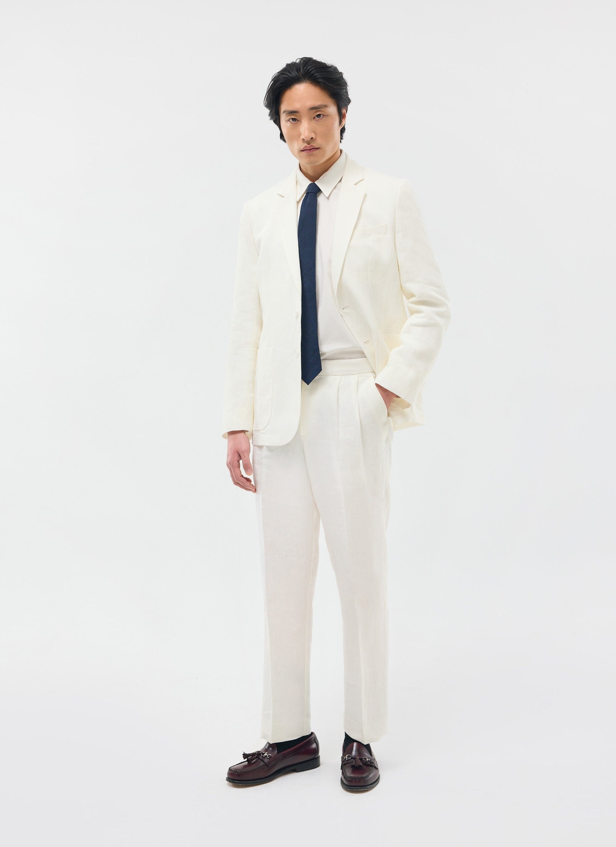 Model wears the Percival Relaxed Tailored Linen Blazer in white with matching trousers, a navy tie, and burgundy loafers.