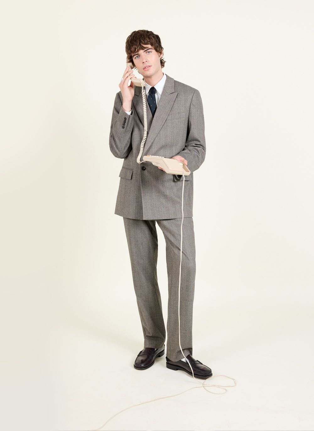 A model wearing the Percival tailored double breasted blazer in grey herringbone with matching trousers on a phone call.