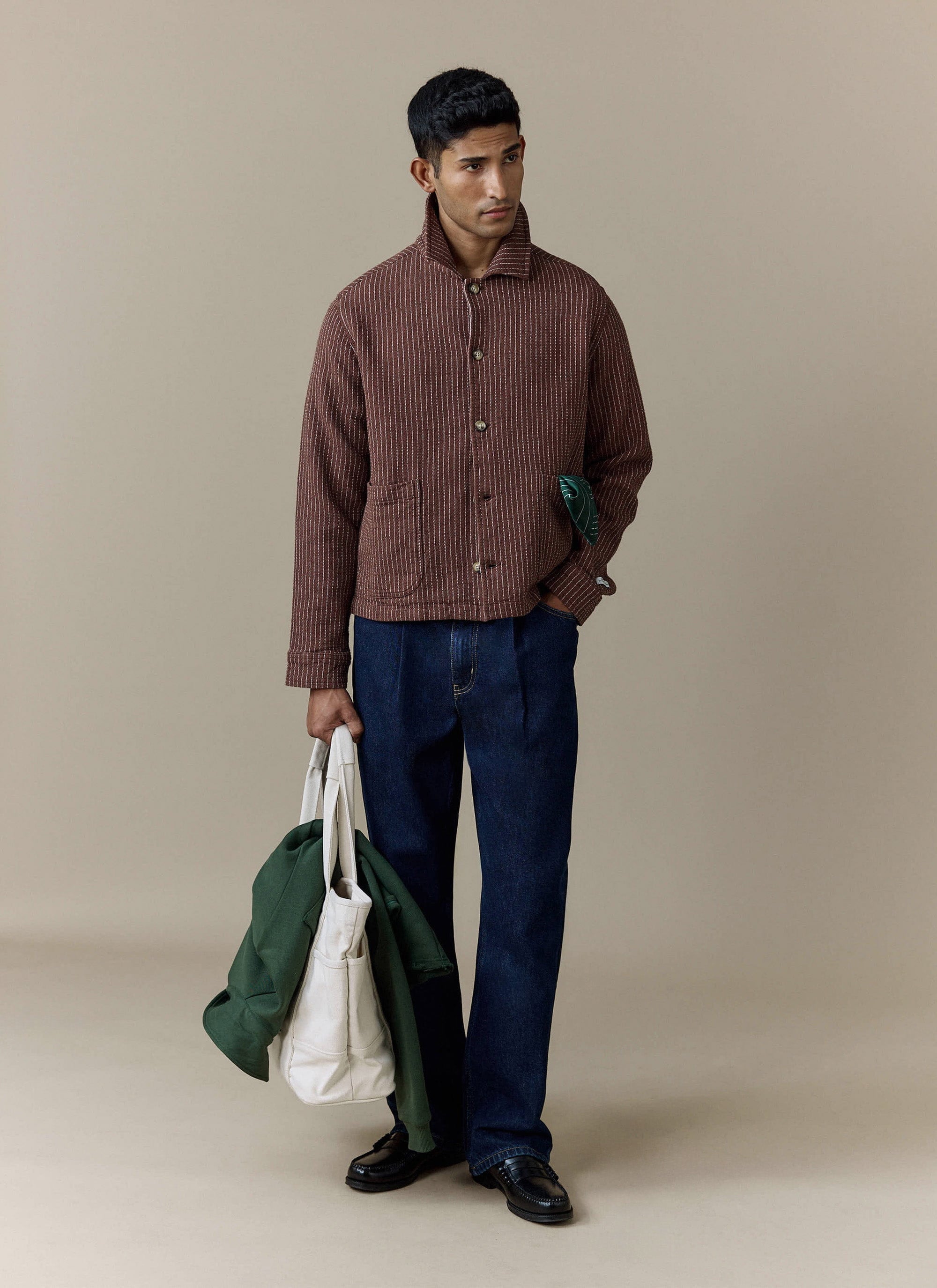 A full-length shot of a model wearing the brown Sorrento Boxy Overshirt from Percival with pleated dark wash jeans.