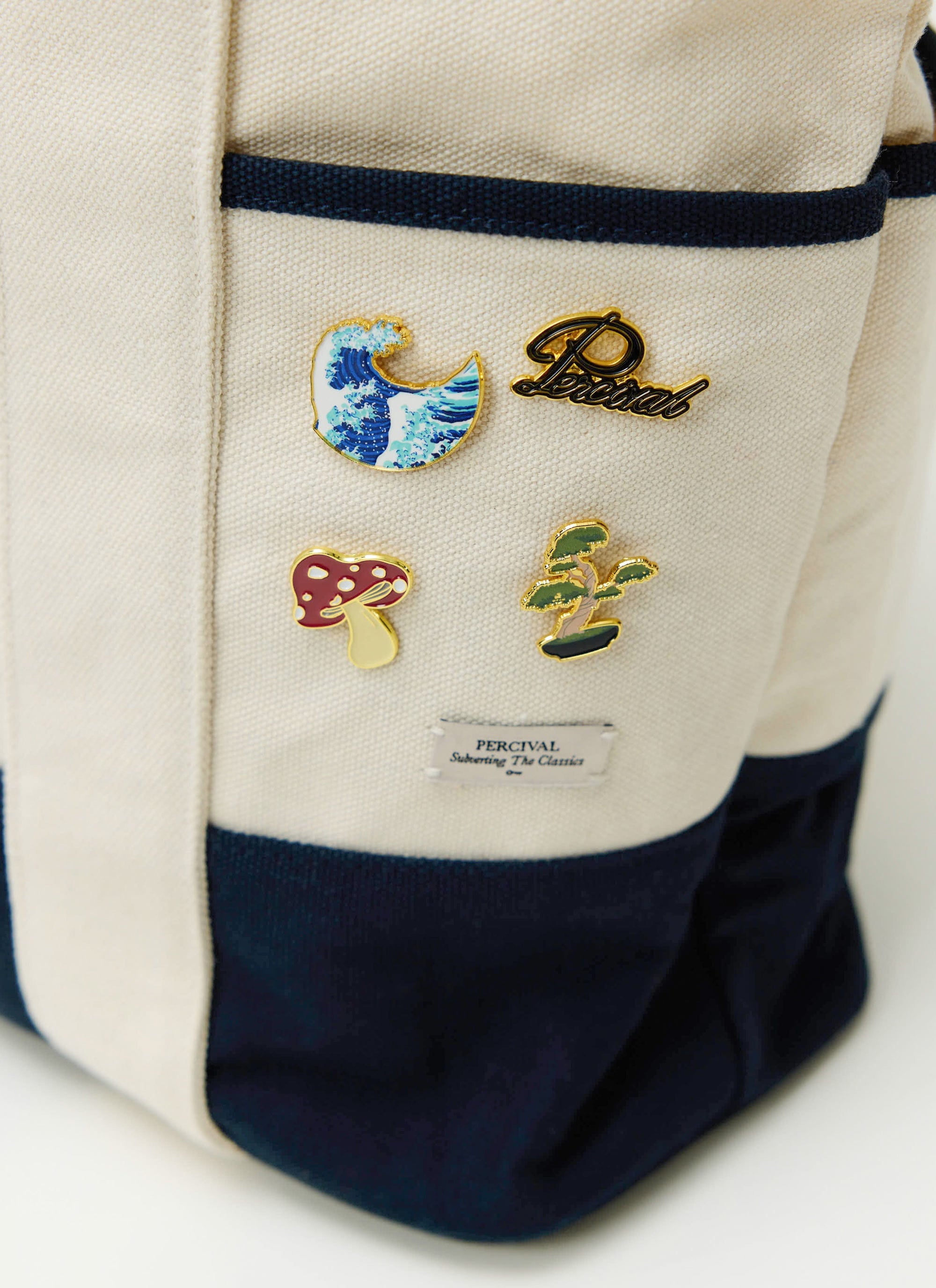 A collection of Percival pin badges, including a mushroom, wave, and bonsai tree, pinned to a canvas tote bag.