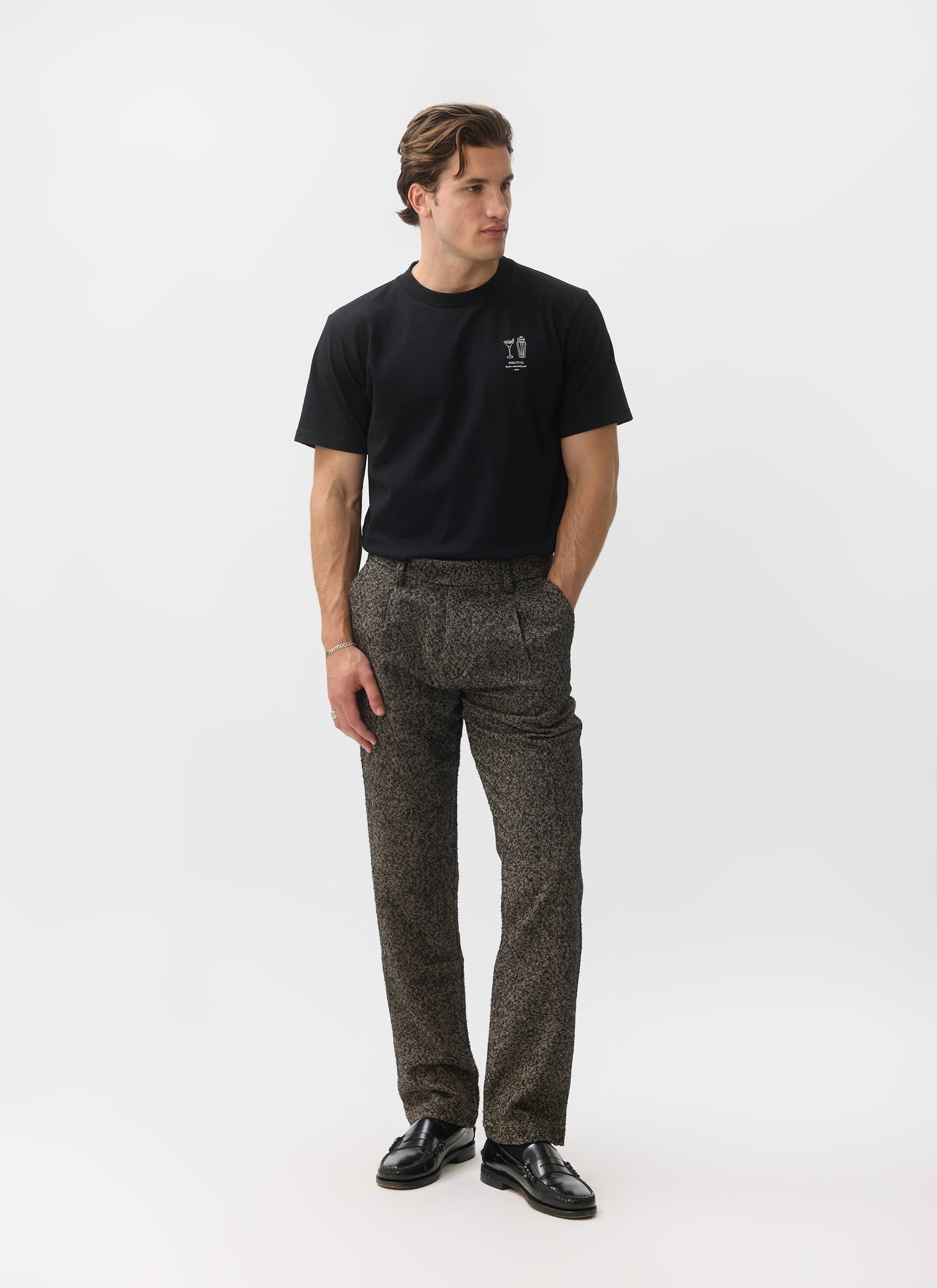 A man models the Percival Blyton Trousers in a warm grey nep fabric against a white studio background.