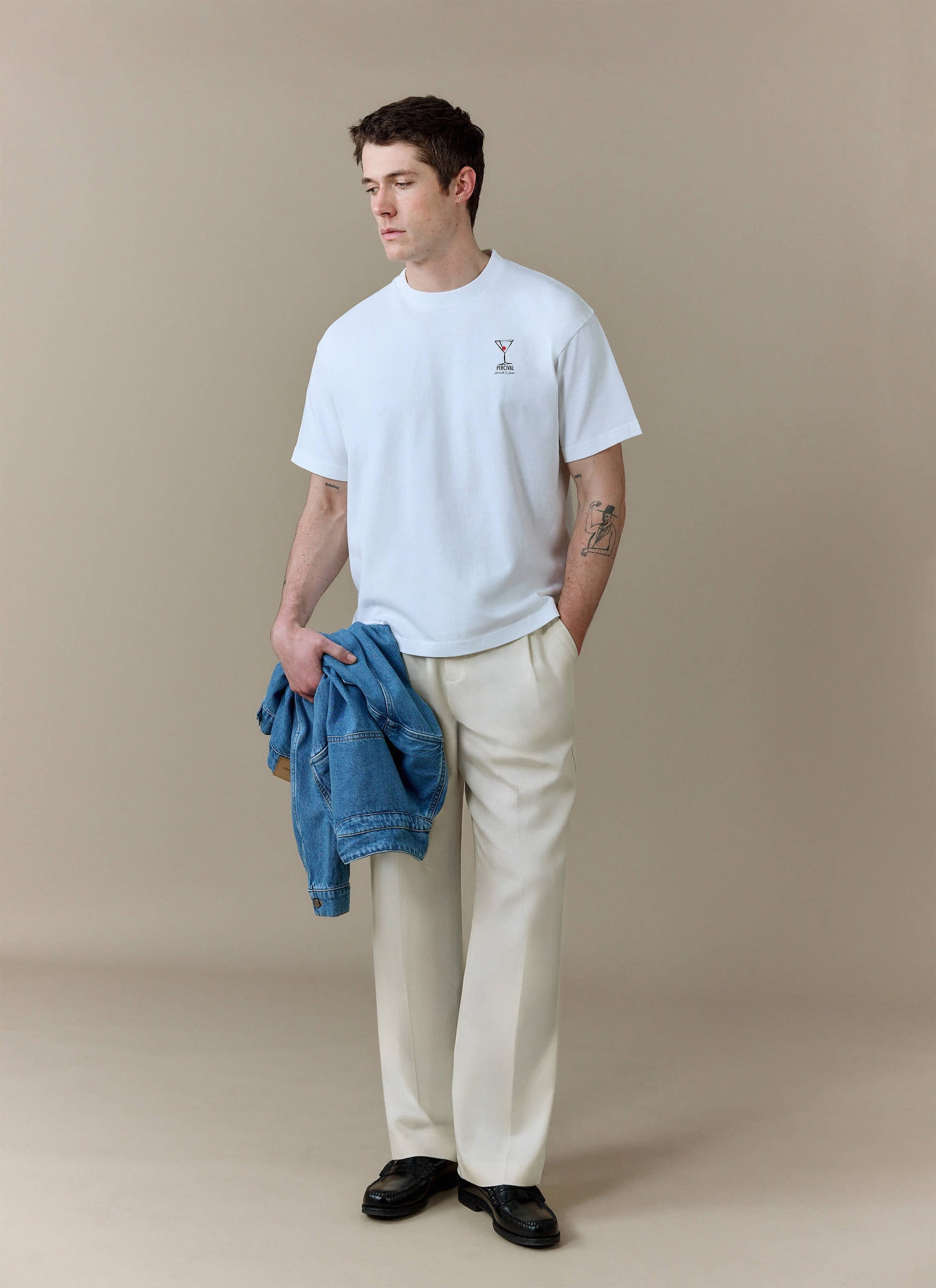 A man models the white Perci and Rossi Martini oversized t-shirt from Percival, styled with cream trousers and black loafers.