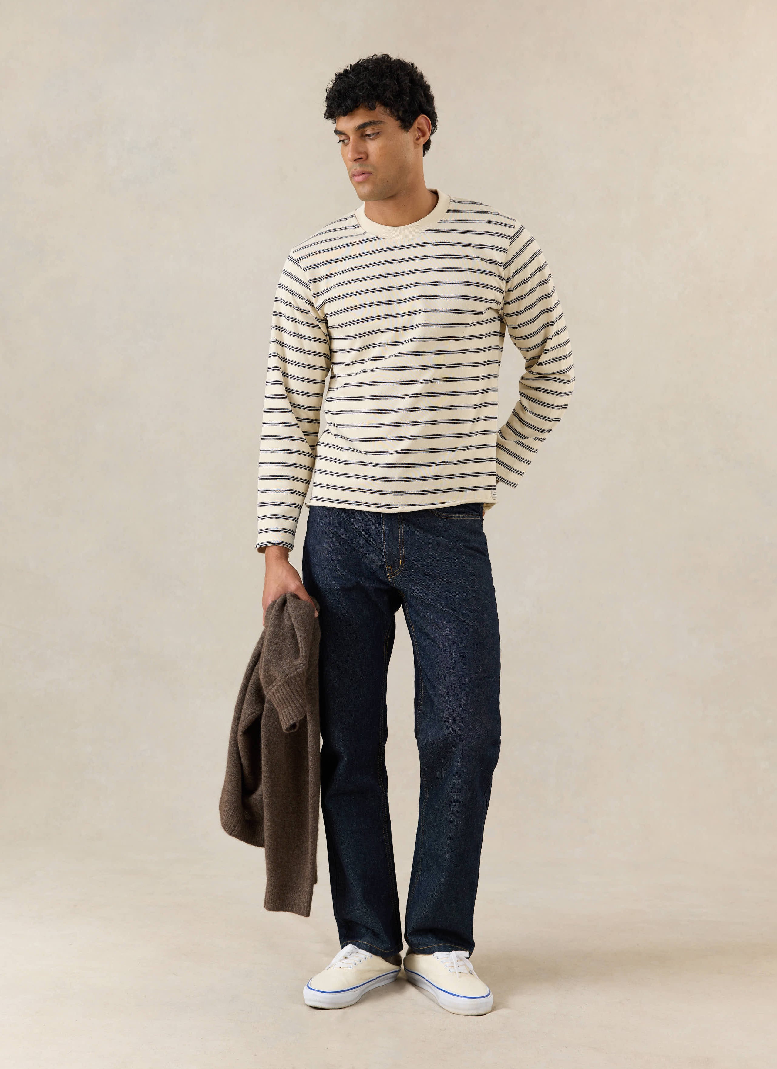 A model wears the Percival Arlo long sleeve Breton T-shirt in white stripe, with dark jeans and a brown jumper.