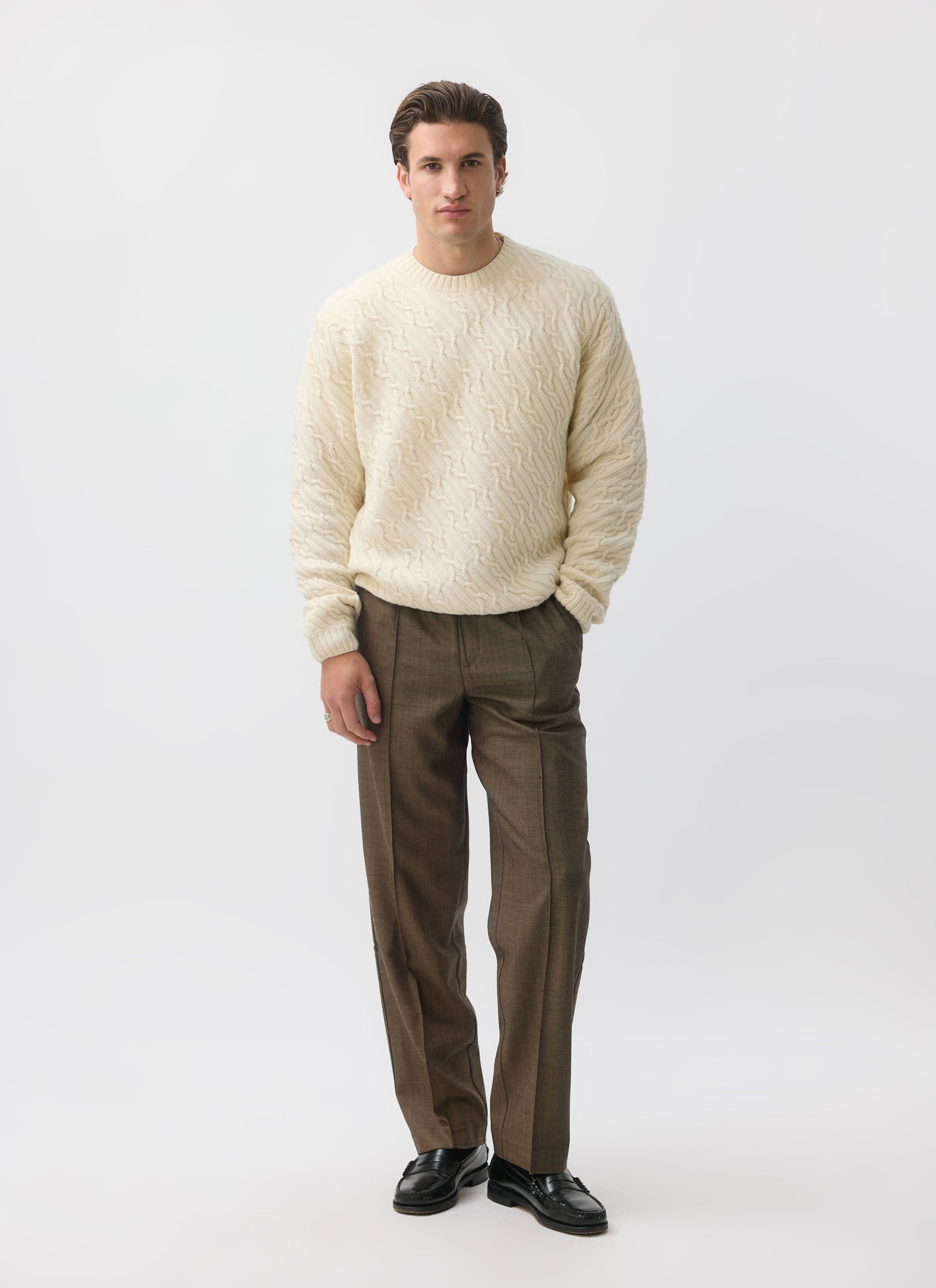 A model wears the Percival Melville Cable Knit Jumper in ecru, paired with smart brown trousers and black loafers.