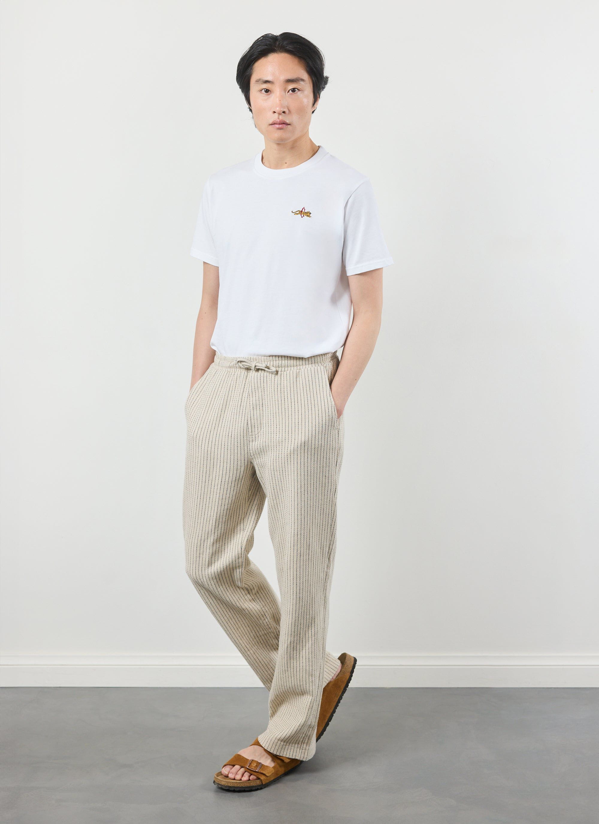 A man models the Percival Sorrento Linen Trousers in ecru, paired with a white embroidered t-shirt and brown sandals.