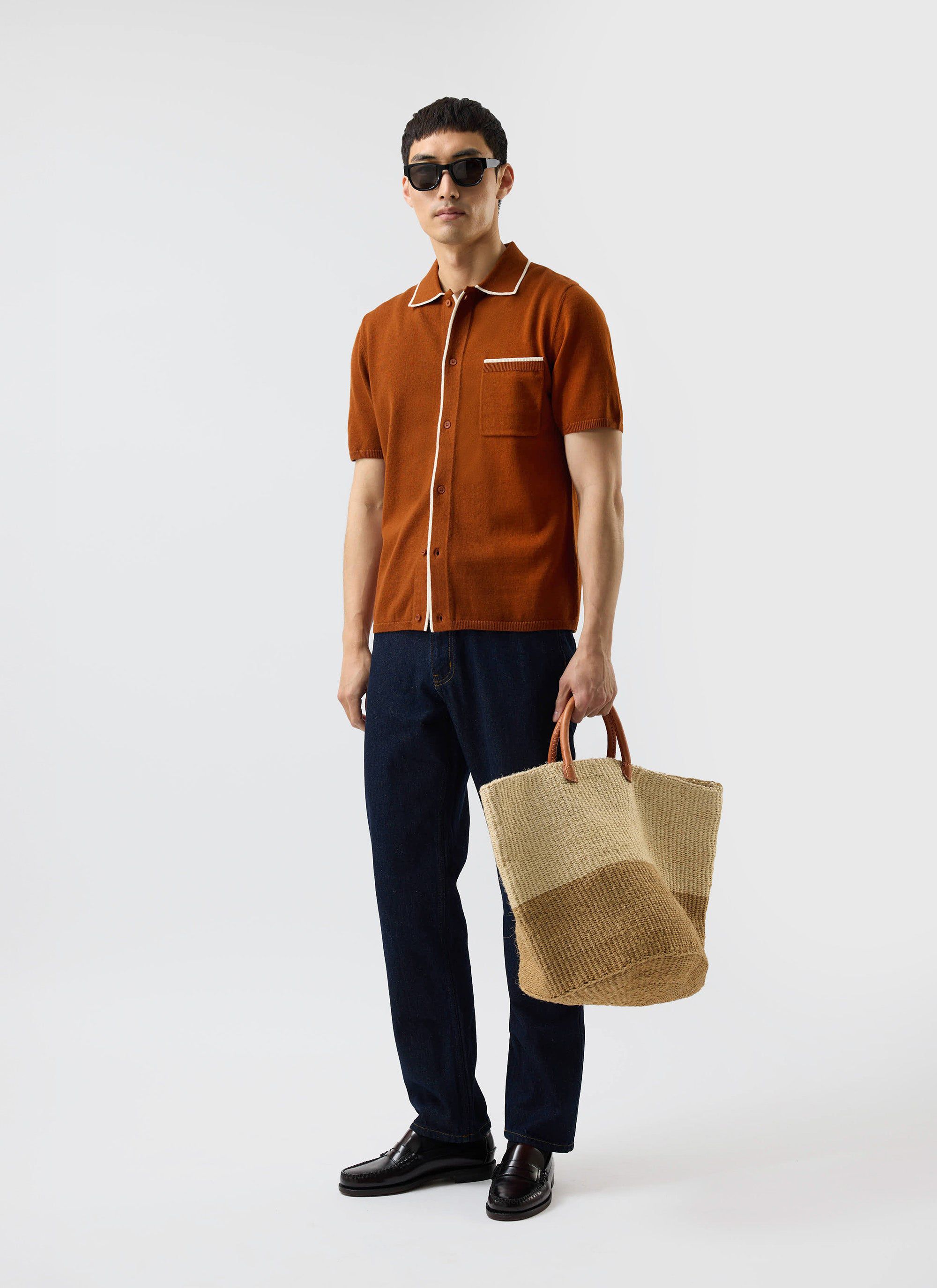 A model wears the Percival Contrast Trim Shirt in rust with dark jeans and sunglasses, holding a woven tote bag.