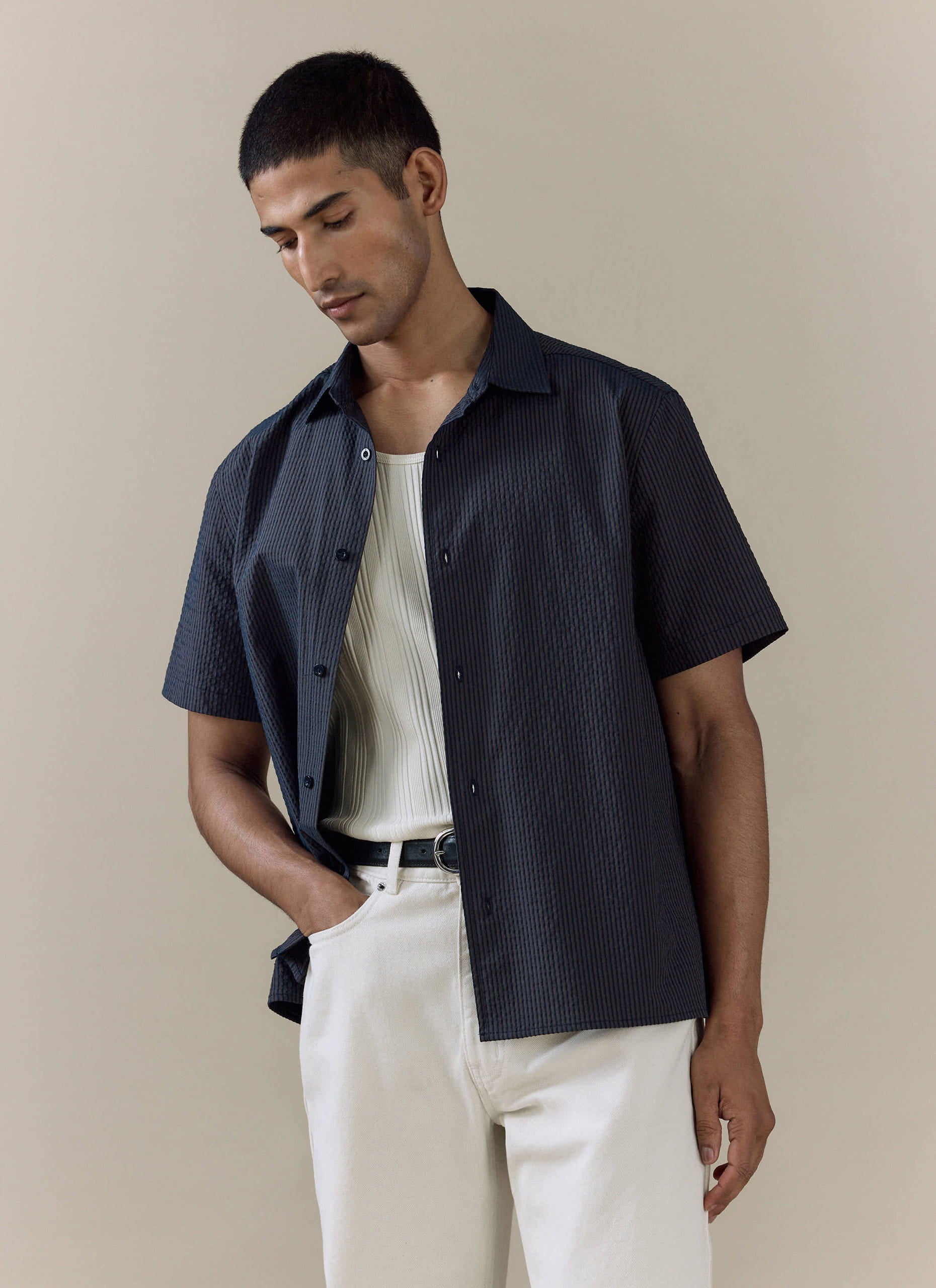 Model wears the navy stripe Rowan Seersucker Boxy Shirt by Percival, styled with a white ribbed vest and trousers.