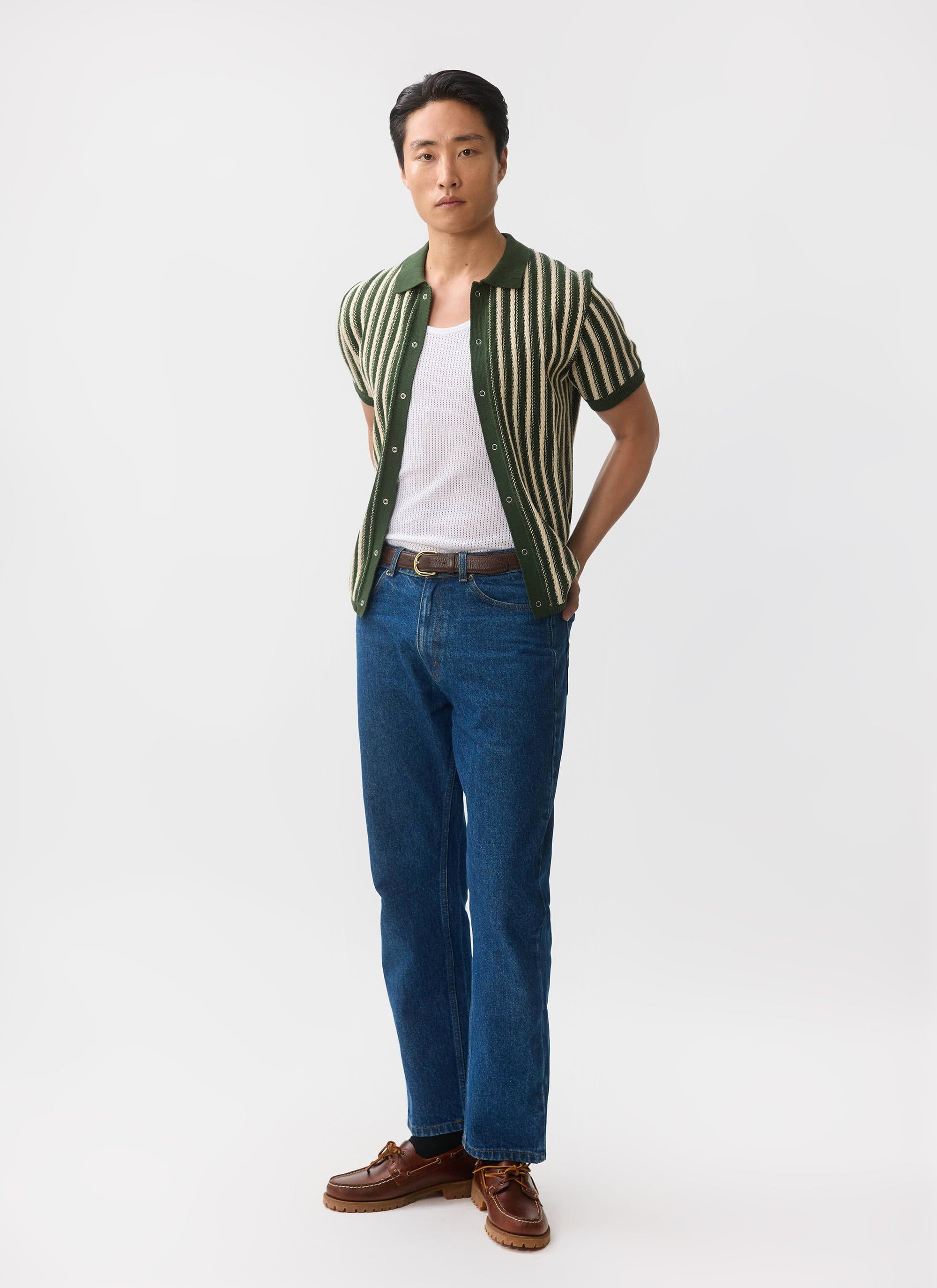 A man models the Percival Collins Micro Cable Knitted Shirt in green, layered over a white vest and styled with blue jeans.