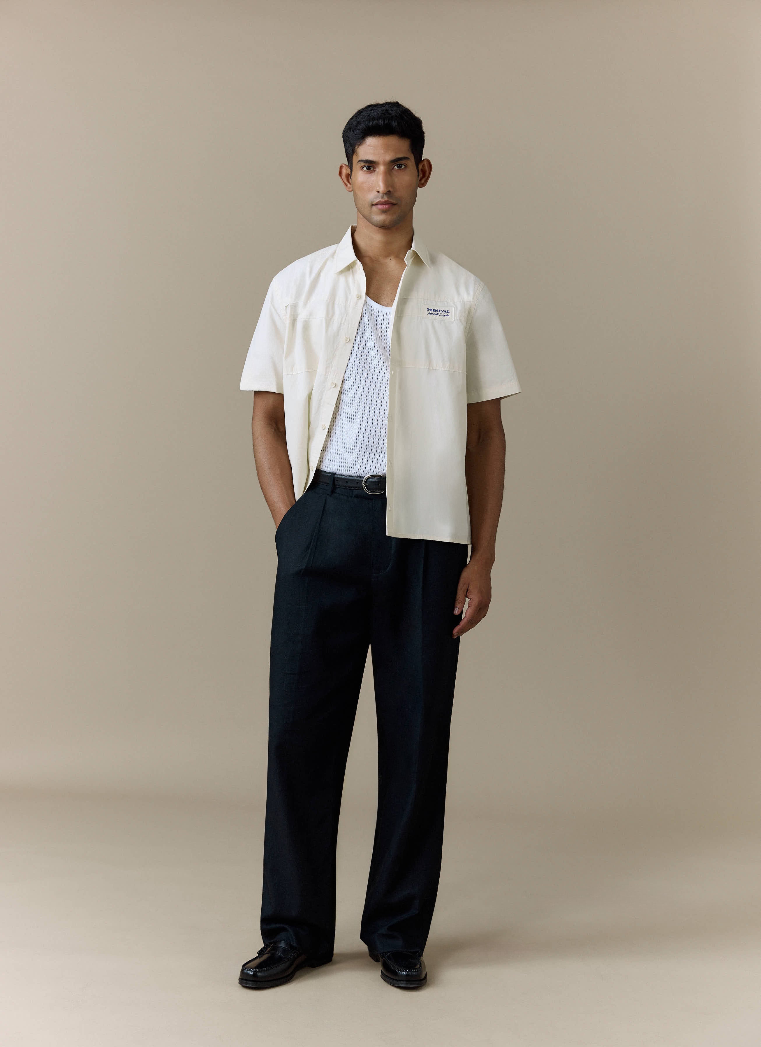 A model wears the Percival Harlow Boxy Shirt in ecru, styled open over a white vest and paired with wide-leg black trousers.
