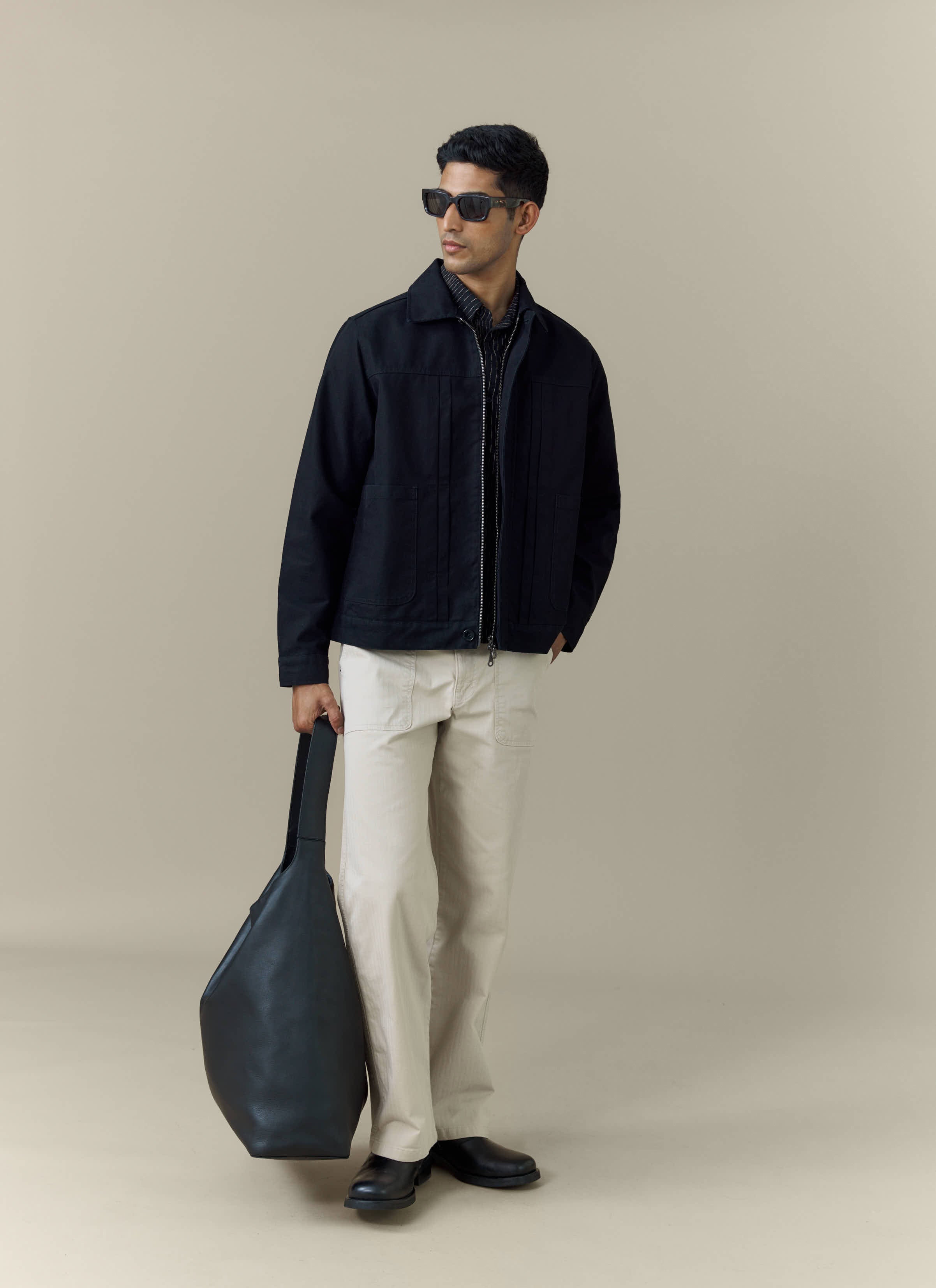 A model wears the black cotton Ernst Jacket from Percival, styled with cream trousers, sunglasses and a black tote bag.