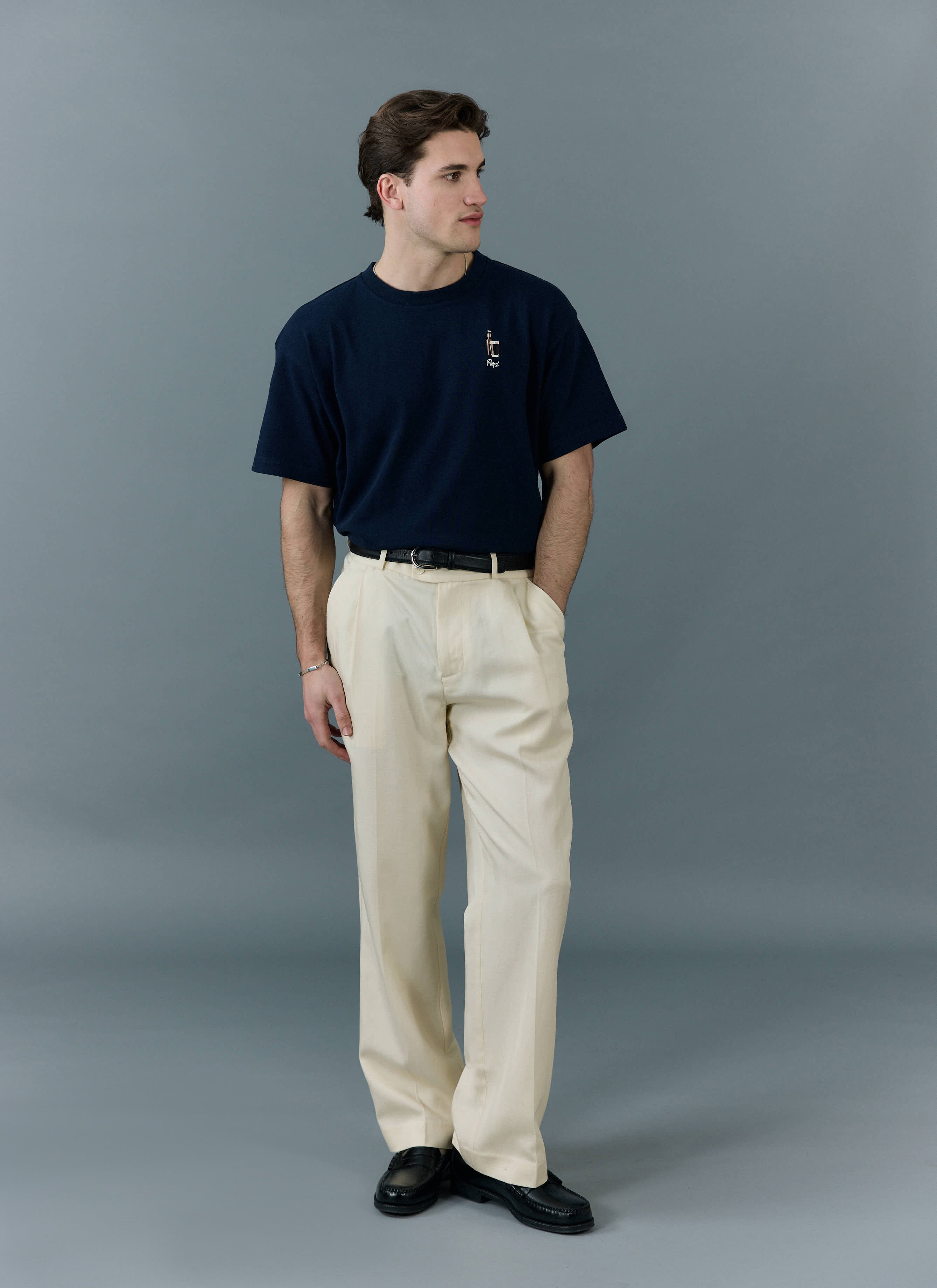 A model wears the navy Percival Stout Oversized T Shirt, styled with cream pleated trousers and black leather loafers.