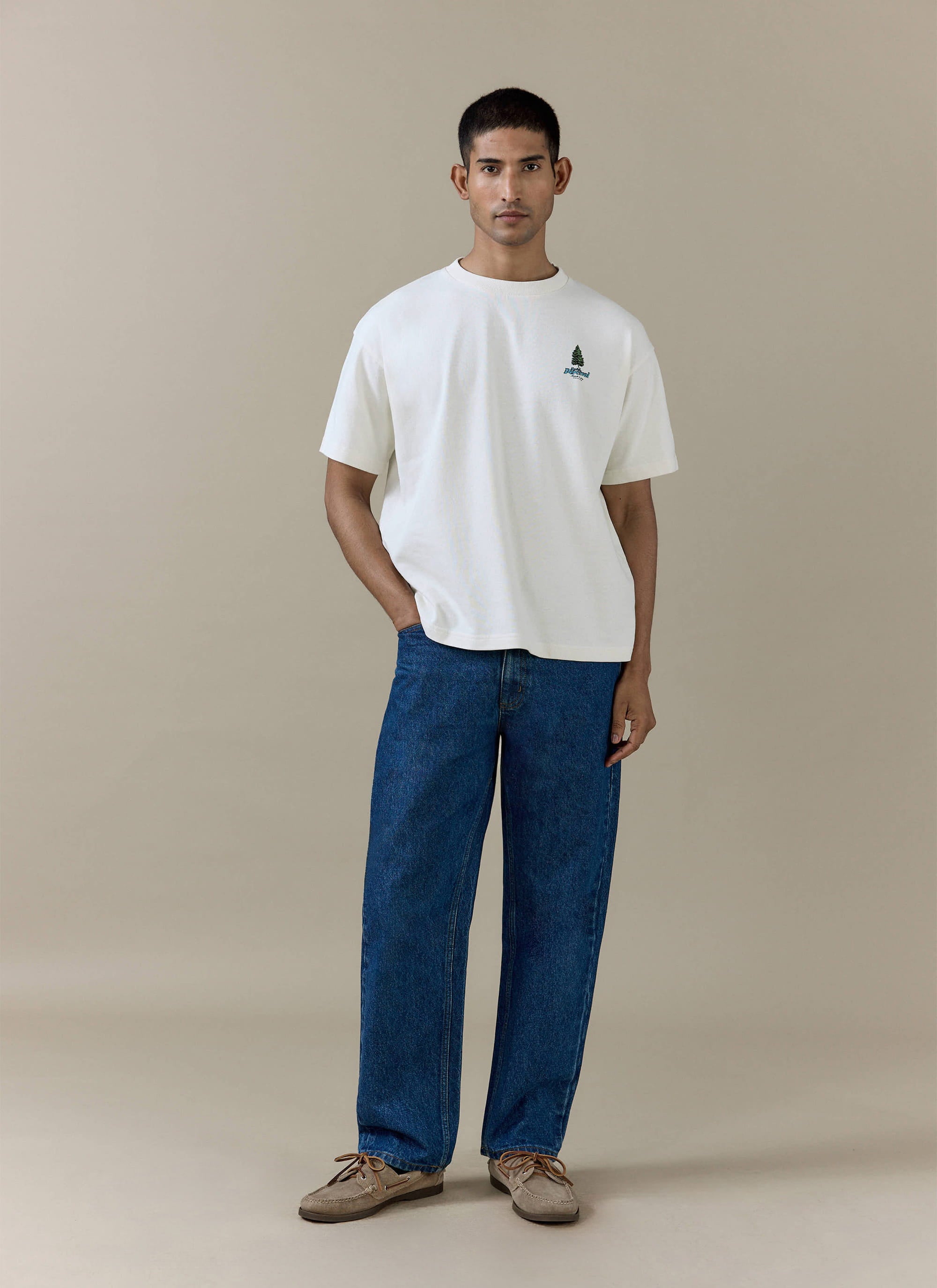 Percival Arnold Oversized T Shirt in cream organic cotton with a pine tree chest graphic, worn with dark blue jeans.