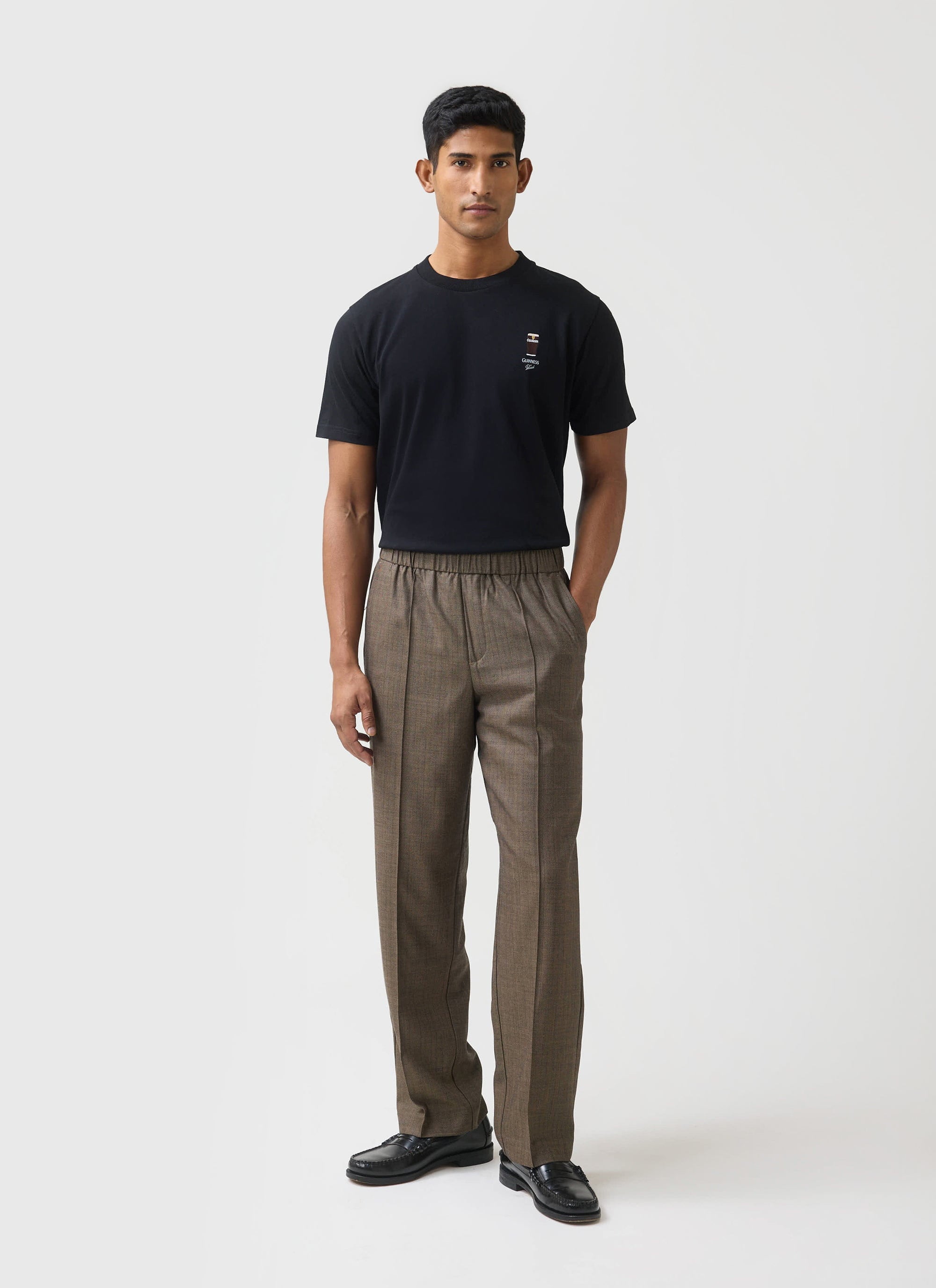 A model wears the black Percival x Guinness Stout T-shirt, styled with smart brown checked trousers in a studio.