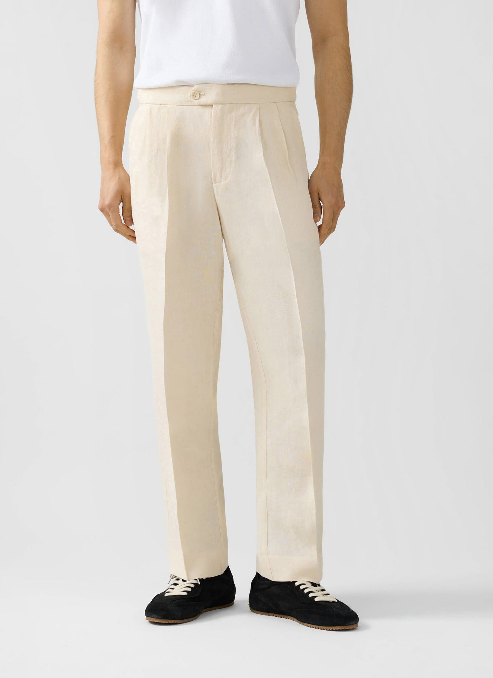 Man wearing Percival cream pleated linen trousers with a white t-shirt and black trainers against a white background.