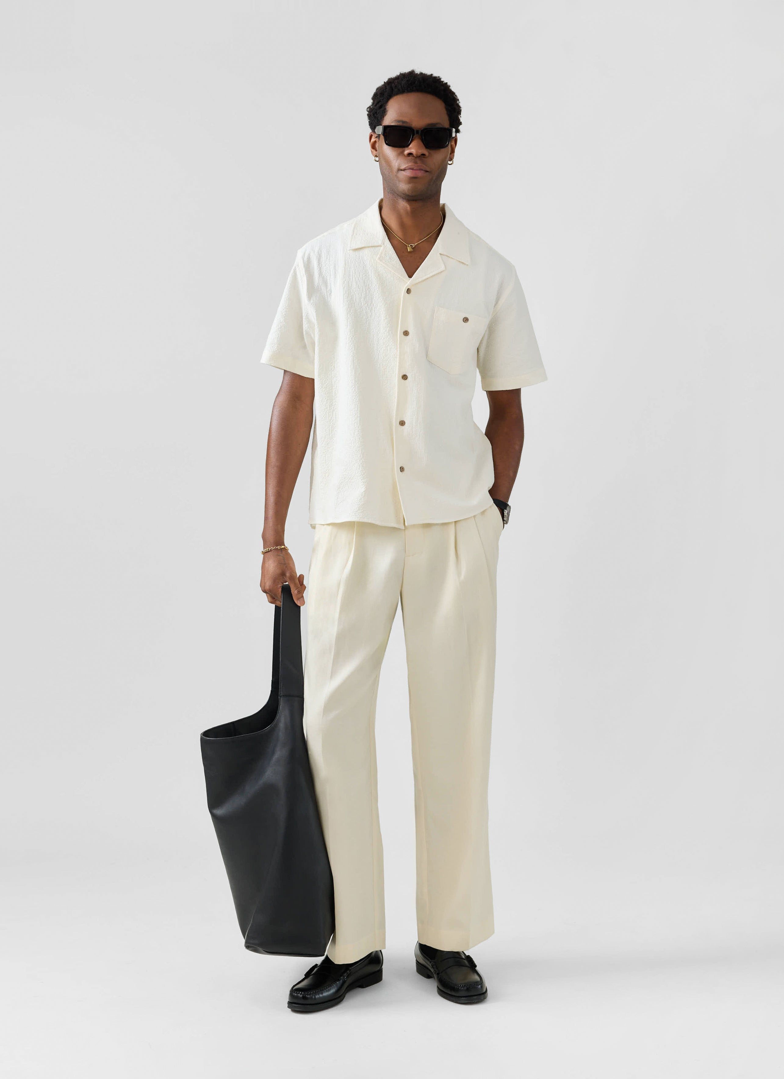 A model wears the Percival ecru seersucker Cuban shirt, styled with matching trousers, sunglasses and a black tote bag.