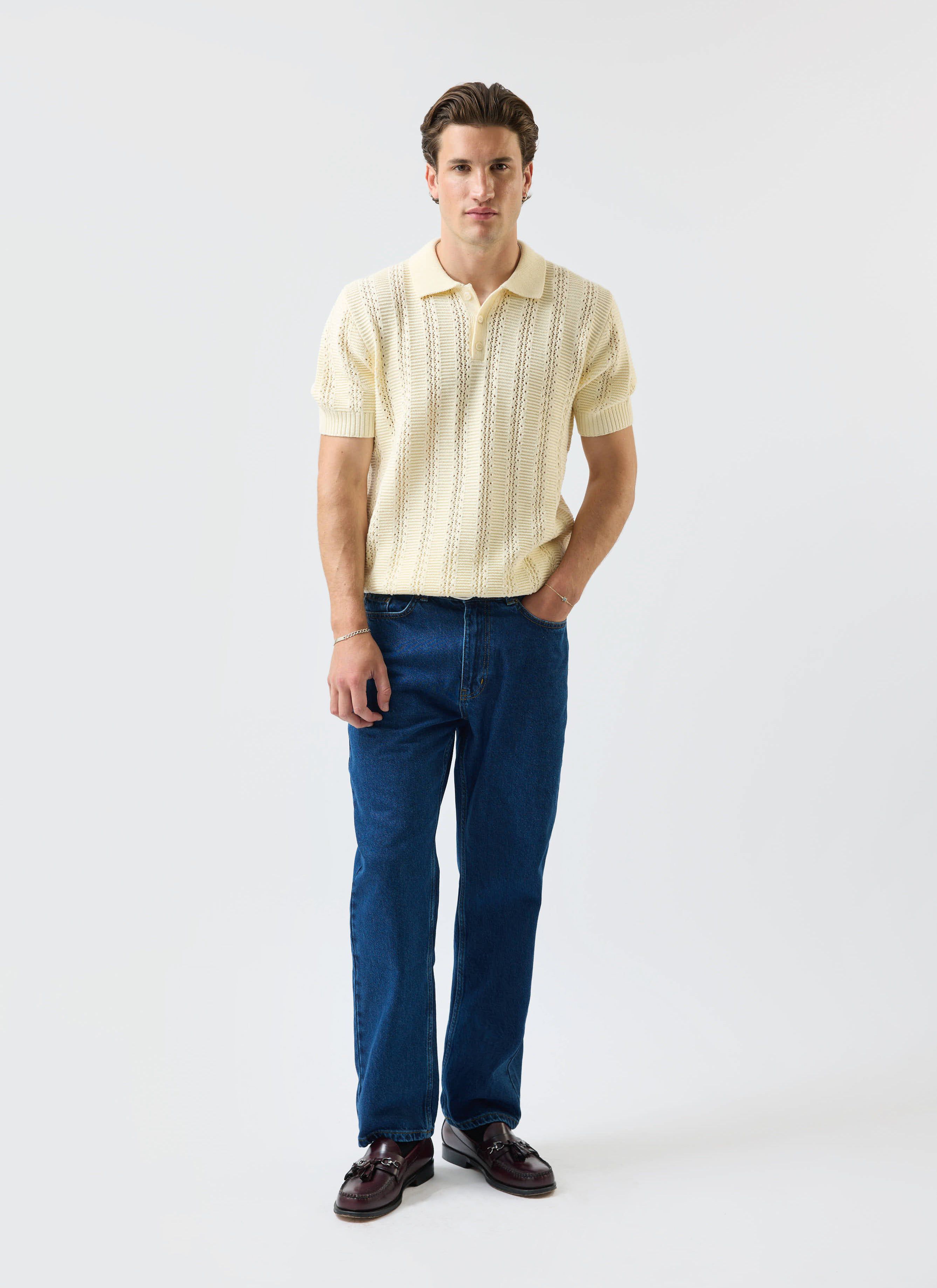 A model wears the Percival Addendum Knitted Polo Shirt in ecru cream, styled with dark blue jeans and loafers.
