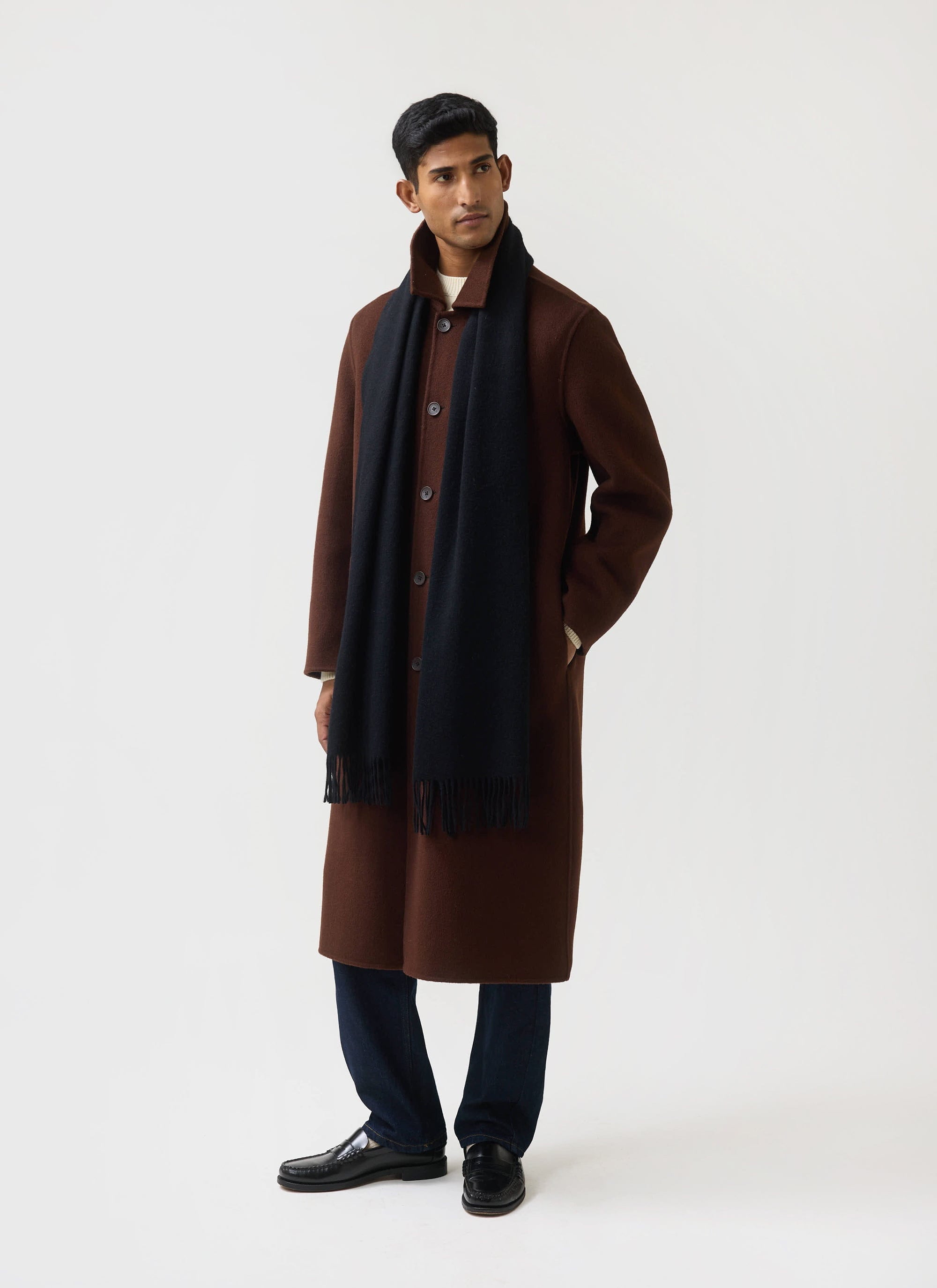A model wears the black wool Tolkein Scarf from Percival, styled with a long brown coat against a white studio background.