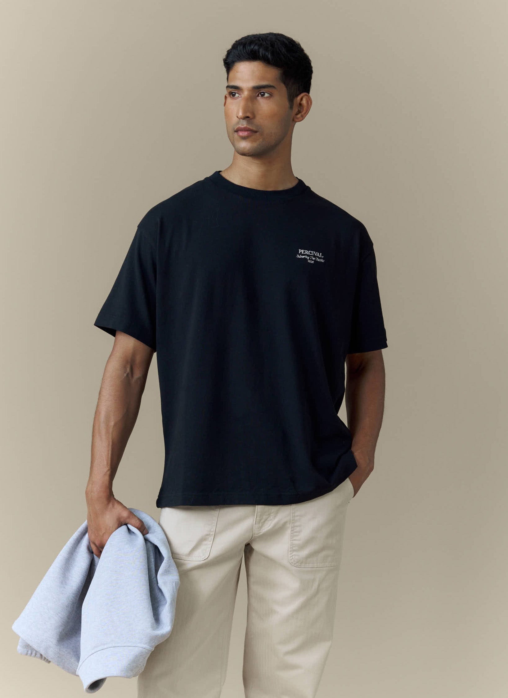 A model wears the Percival Perci Fruit Farm oversized t-shirt in black, styled with cream trousers and holding a sweatshirt.
