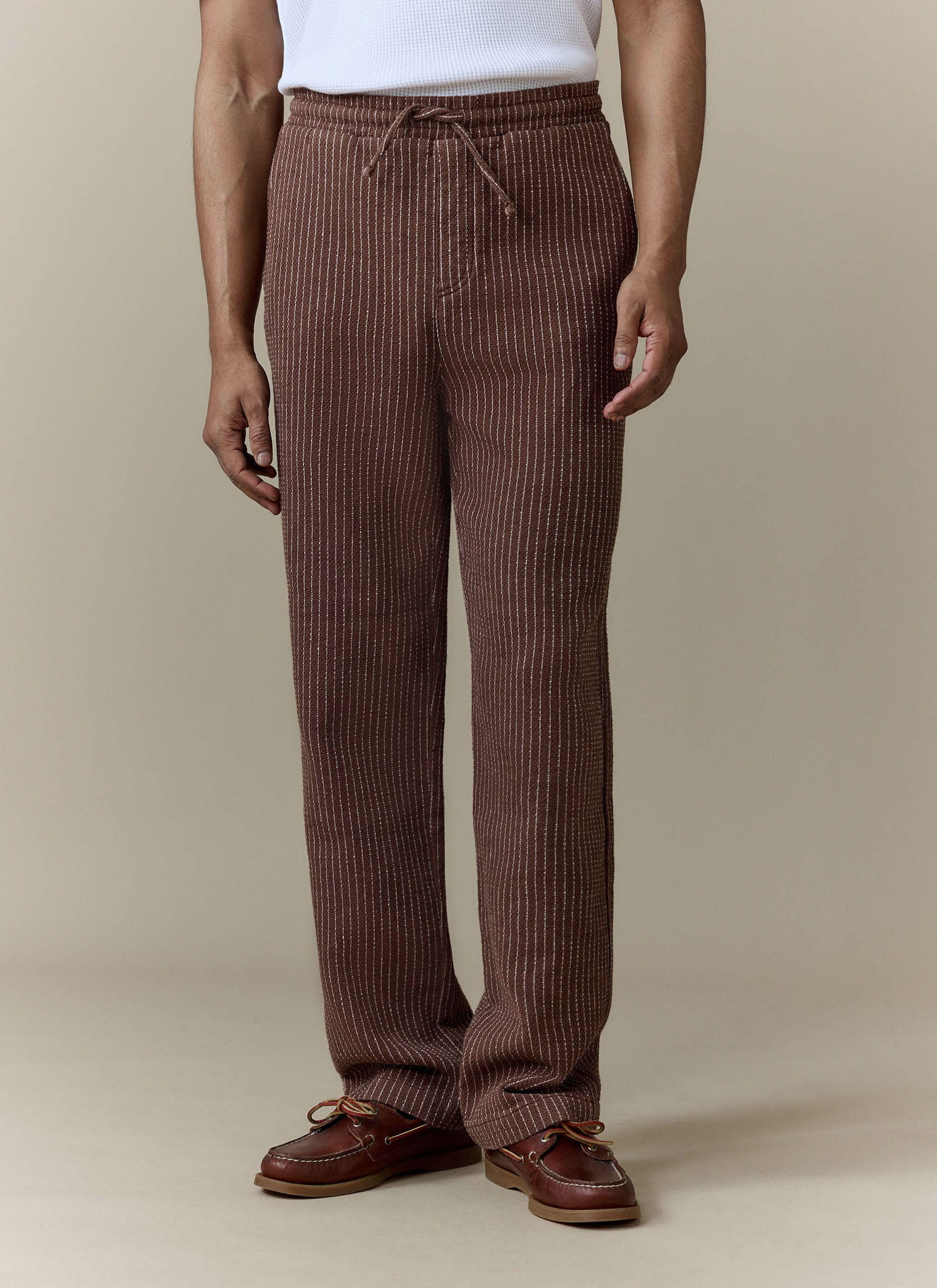 A model wears the brown pinstripe Sorrento Wide Leg Trousers from Percival, styled with a white top and brown boat shoes.