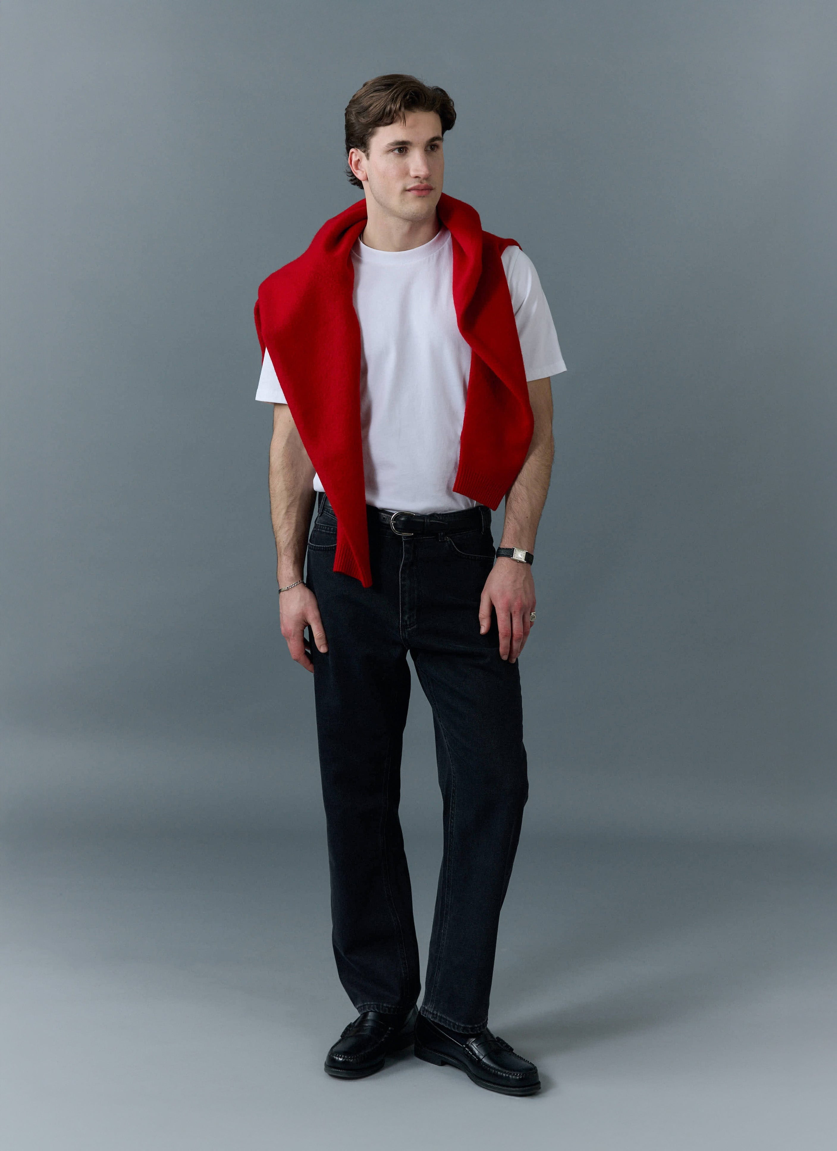 A model wears Percival black straight leg jeans with a white t-shirt and a red jumper draped across his shoulders.