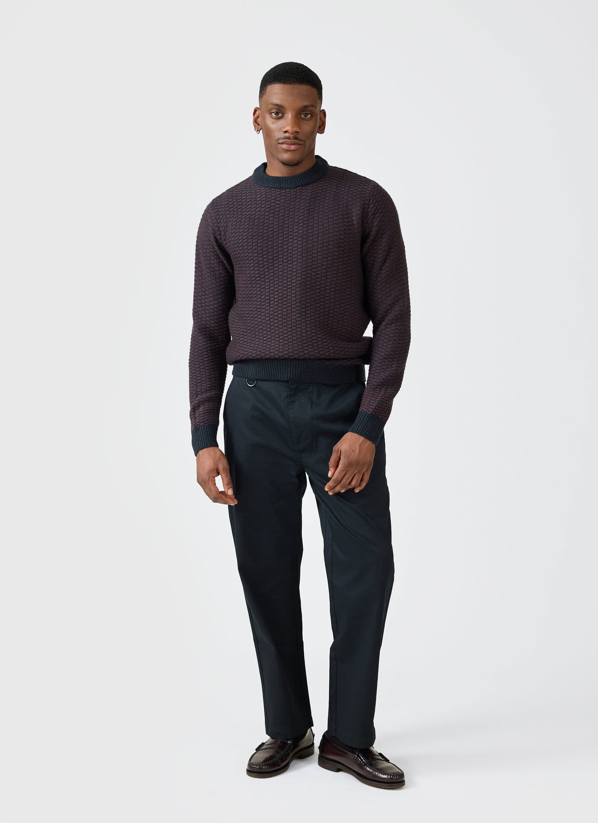 A man models the Percival Charlestown Knitted Jumper in Navy Burgundy, paired with smart black trousers and loafers.