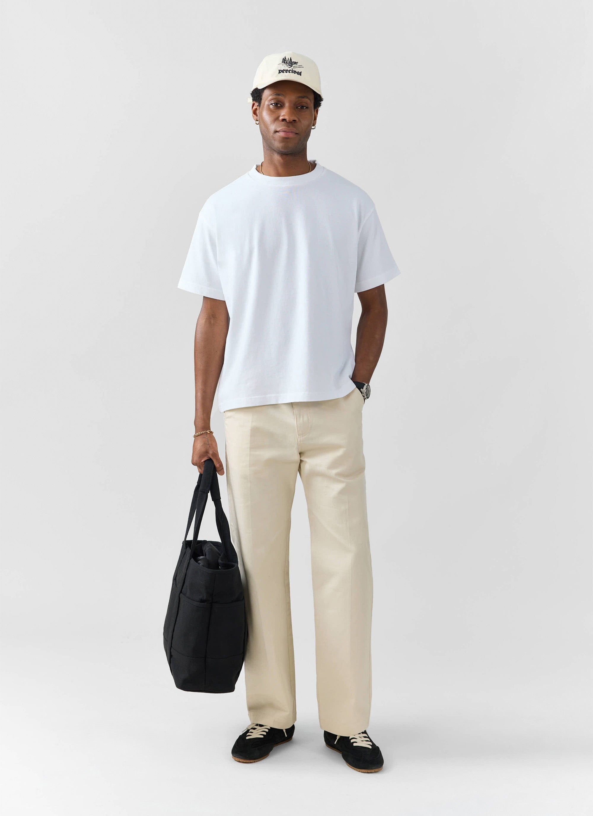 A model wears a white oversized t-shirt from Percival, with cream trousers, black trainers and a cap.