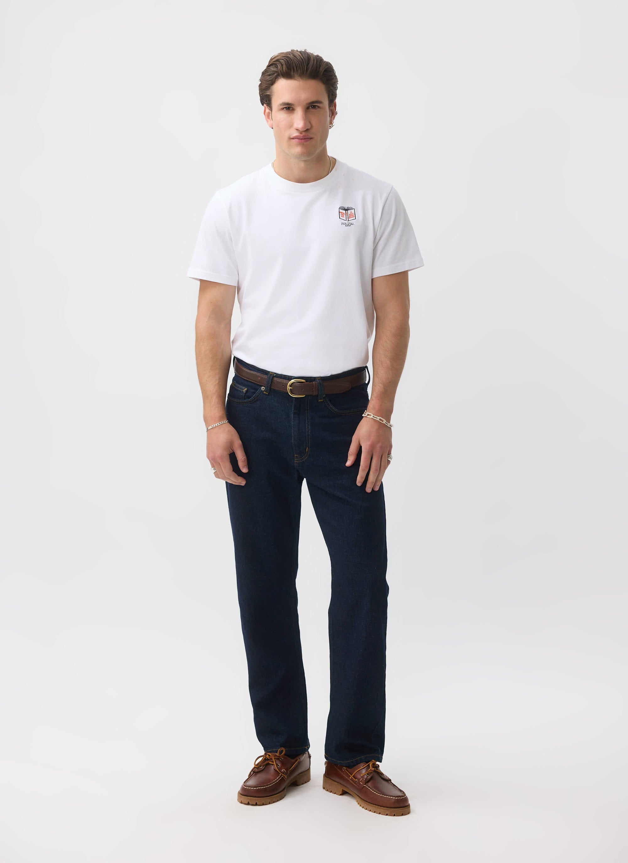 A model wears the white Percival Greatest Bookshop T Shirt, styled with dark wash jeans and brown boat shoes.