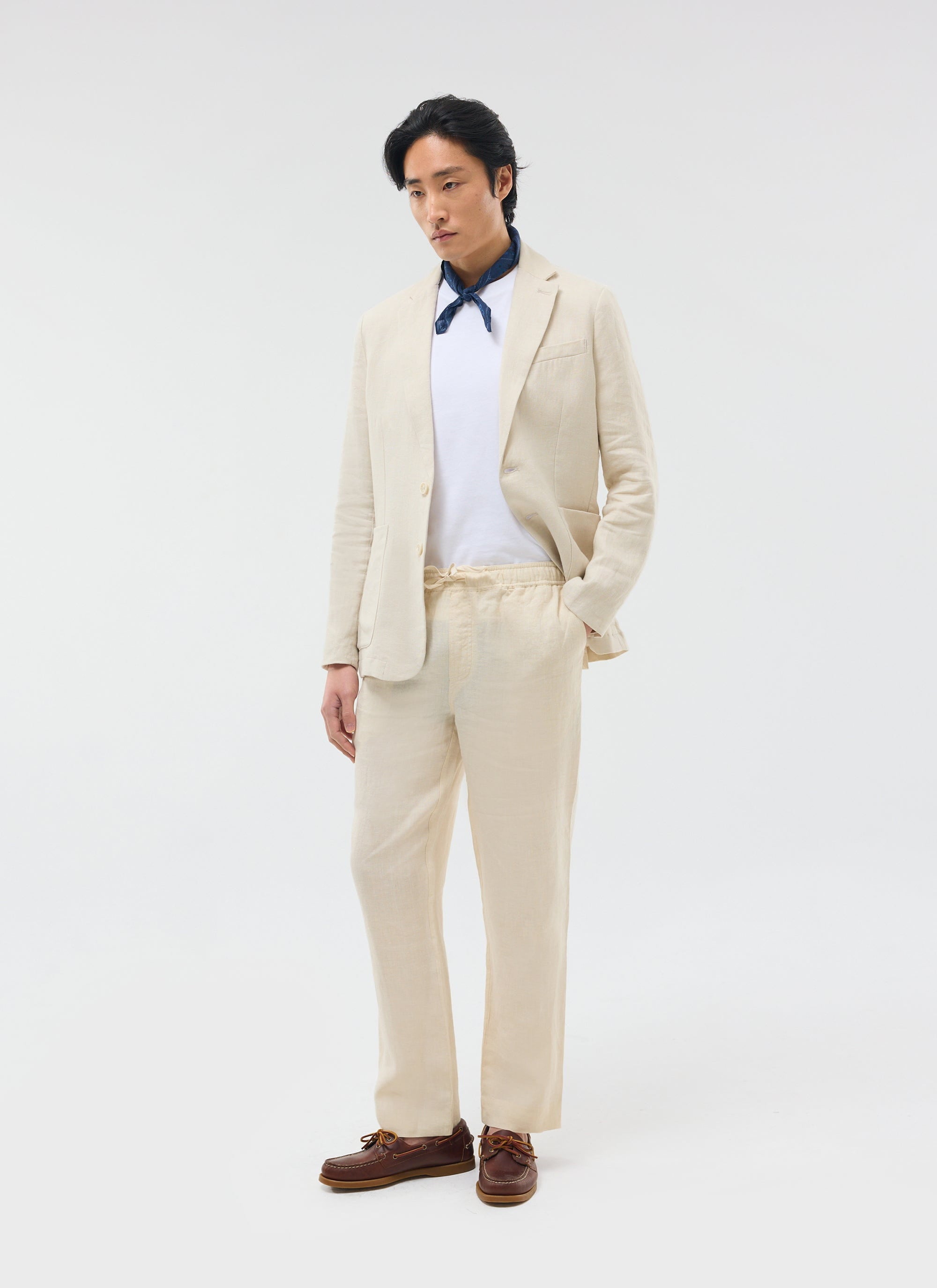 Man wears Percival cream linen trousers and matching blazer with a white t-shirt, blue neckerchief and brown boat shoes.