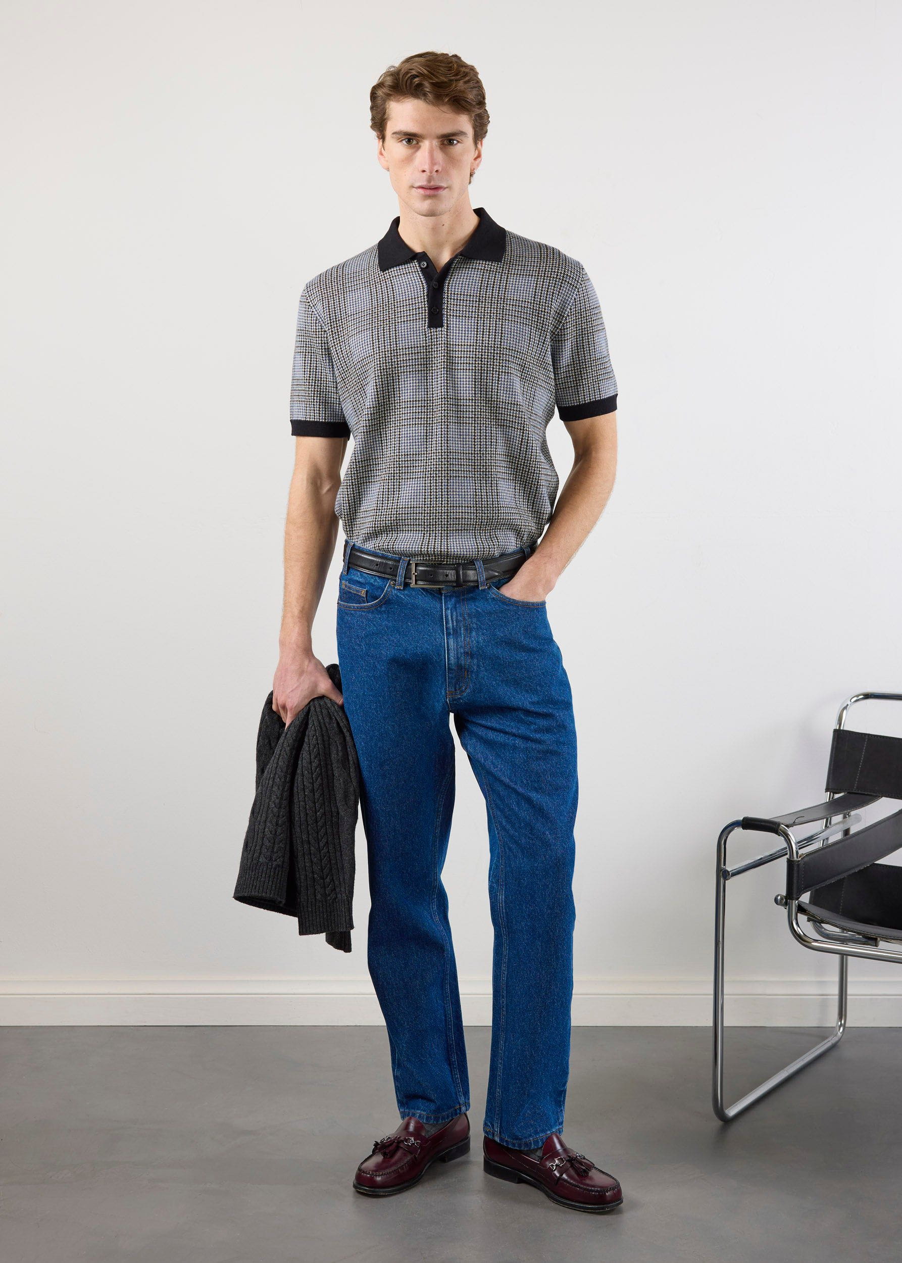 A man models the Percival Casa Martini Knitted Polo in navy puppytooth, styled with blue jeans and loafers in a studio.