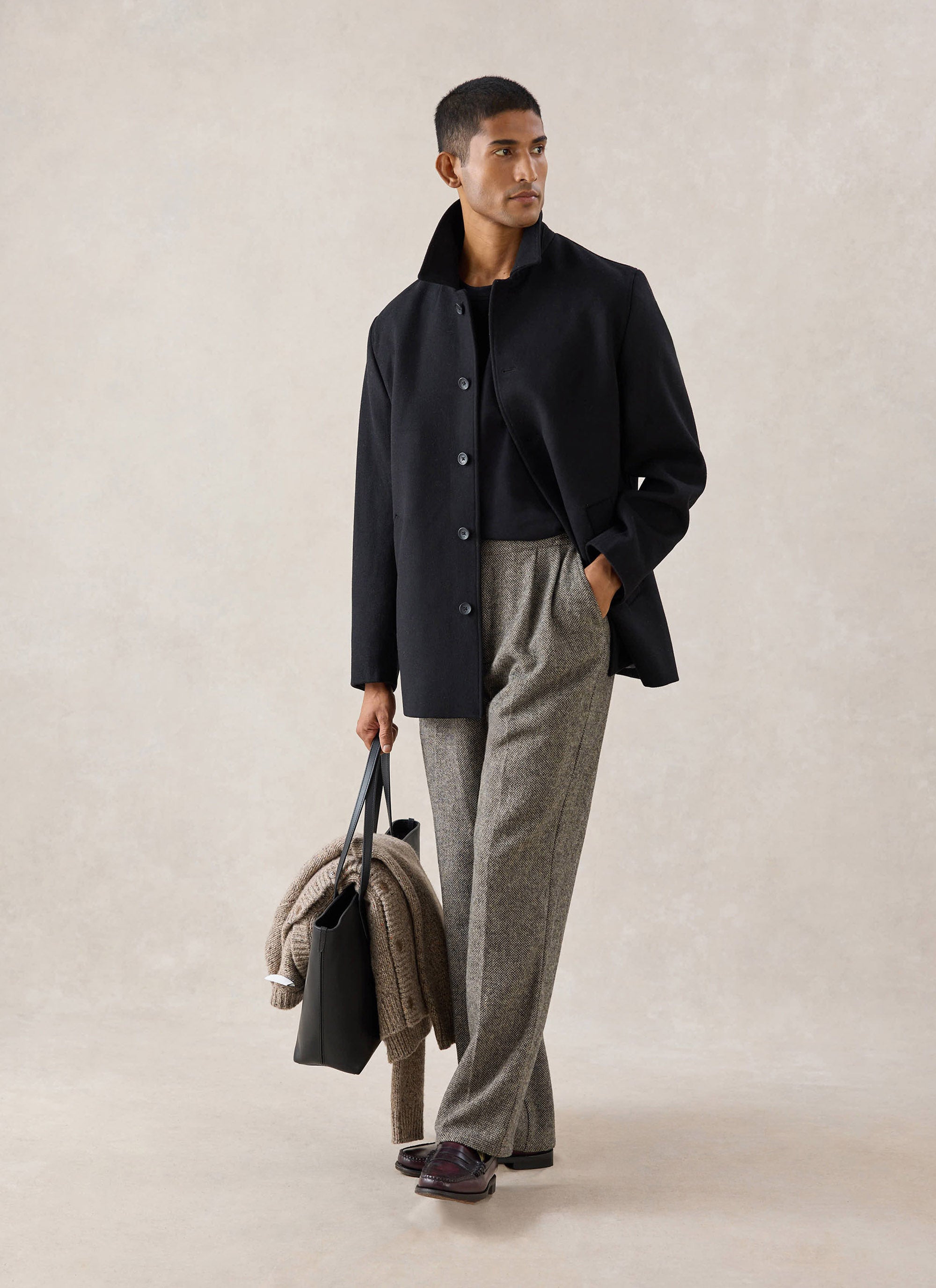 A model wears a black Melton wool peacoat from Percival over patterned trousers, holding a black bag in a studio setting.