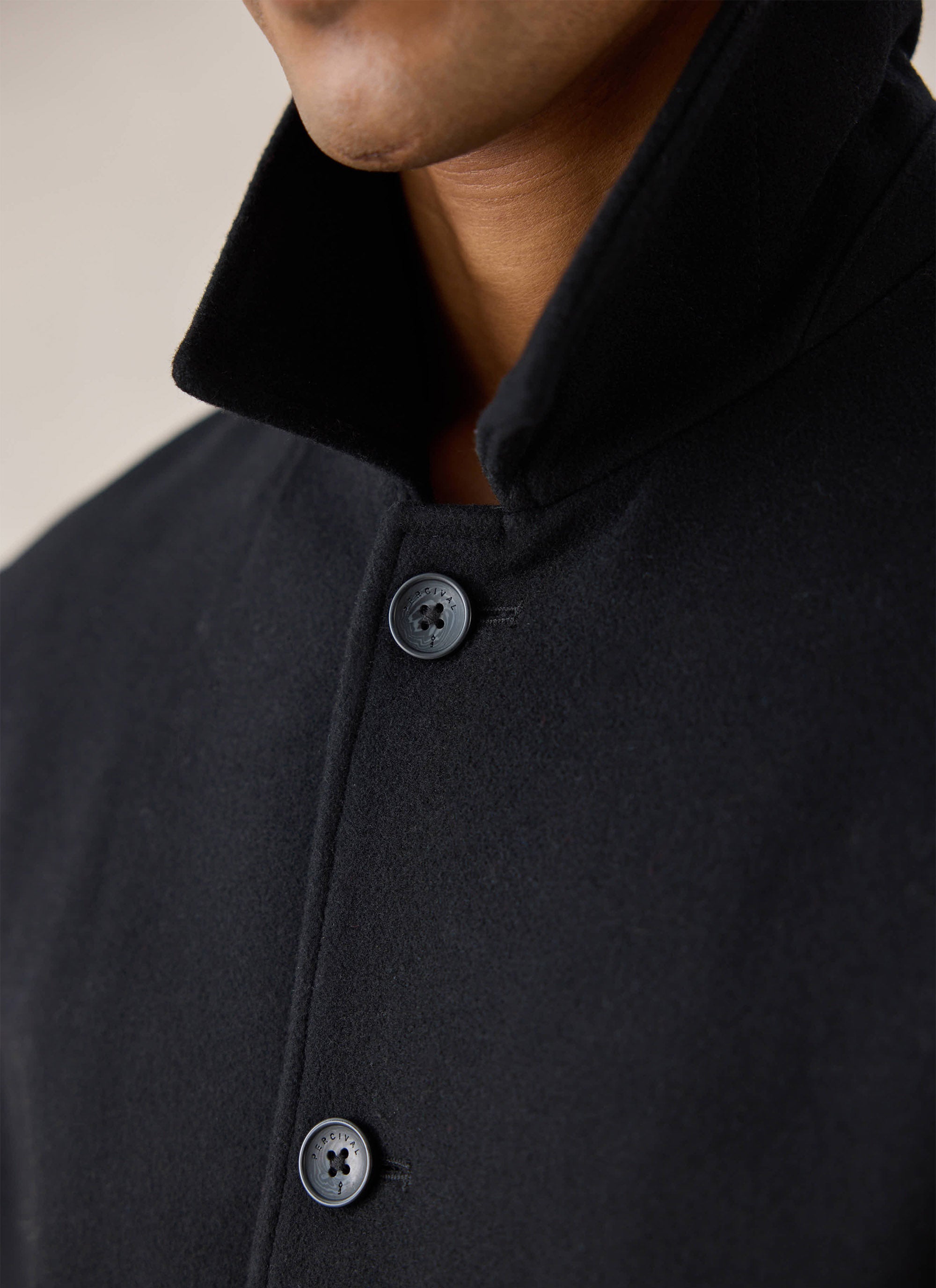 Close-up of the collar and branded buttons on the black Melton wool peacoat from Percival worn by a model.
