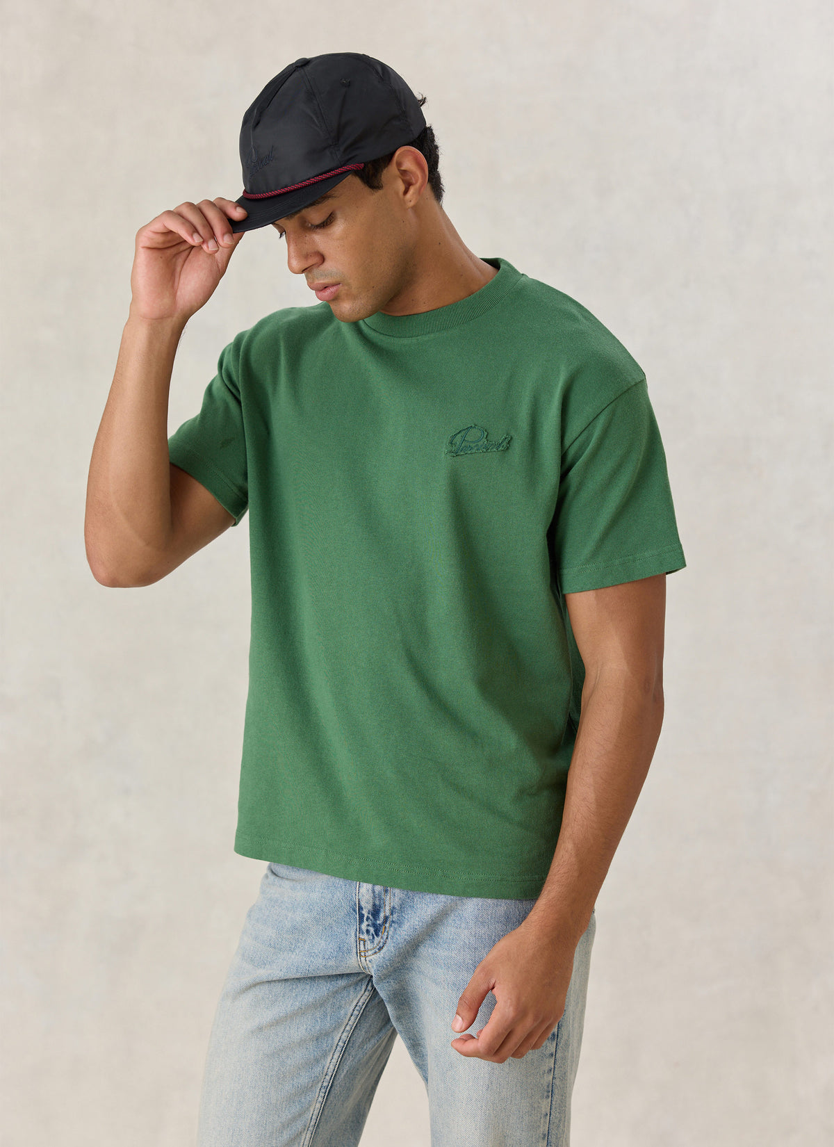 A model wearing the forest green Percival organic cotton oversized T-shirt, styled with a black cap.