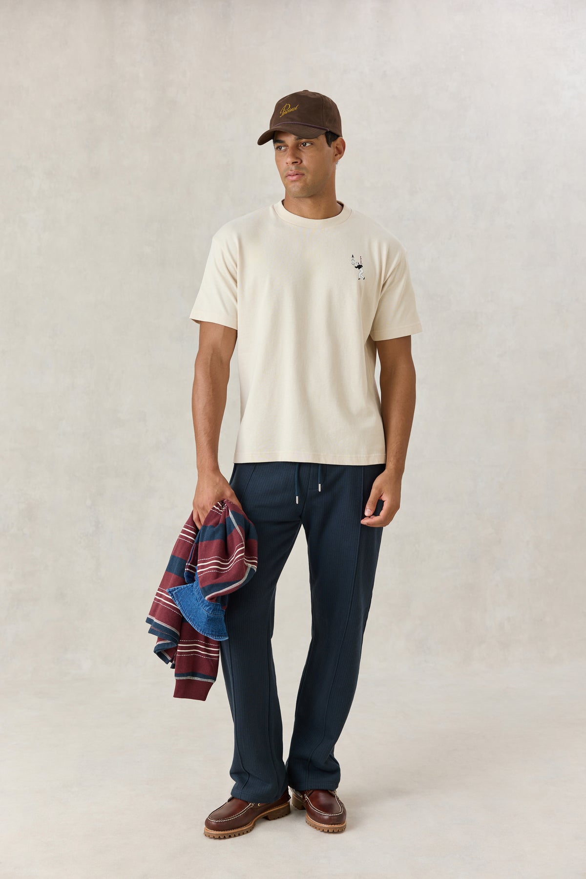 Model wearing the cream Percival oversized T-shirt with waiter graphic, styled with dark trousers, brown cap, and shoes.