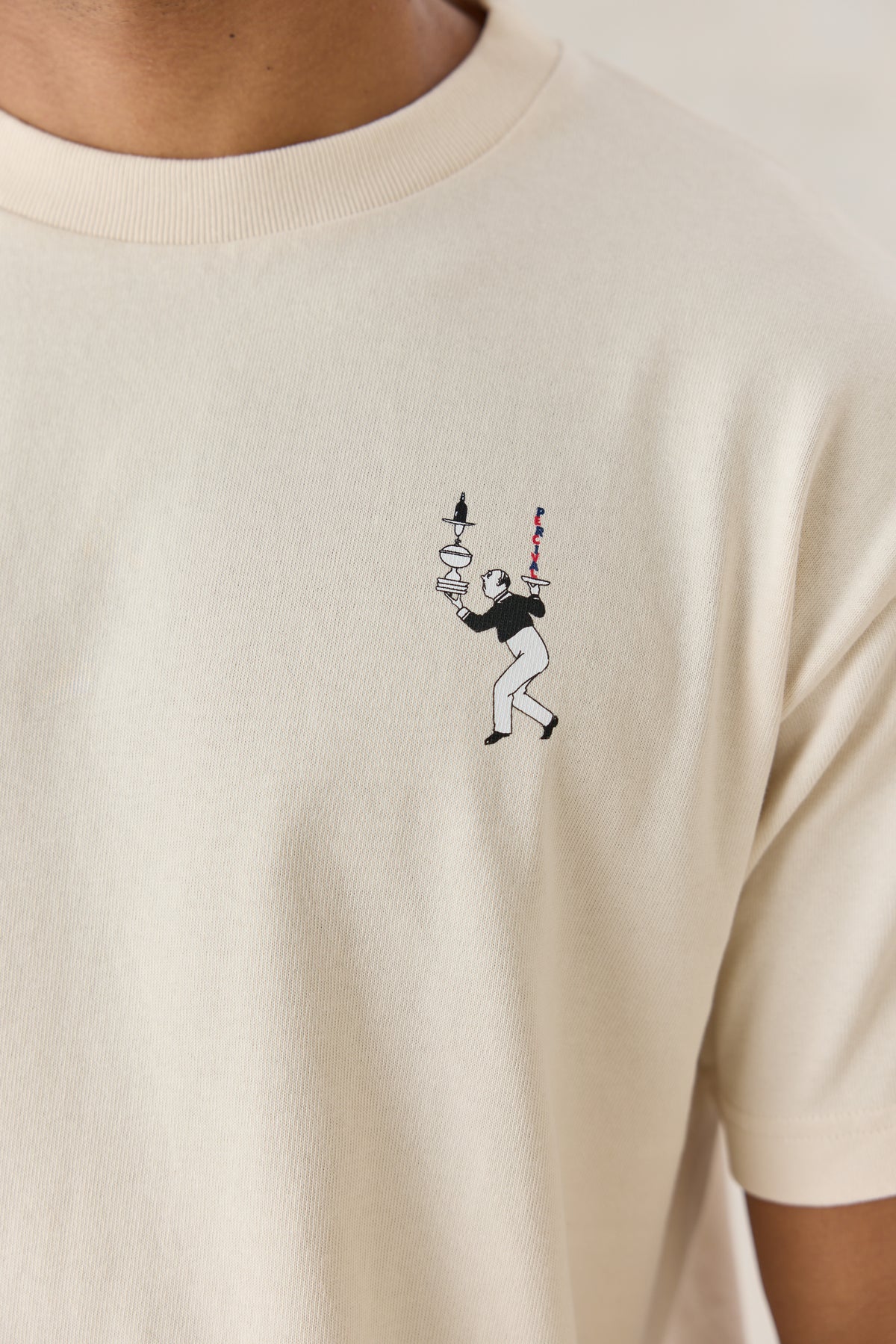 A close-up of the Percival Perci Waiter oversized T-shirt in cream, showing the small waiter graphic at the chest.