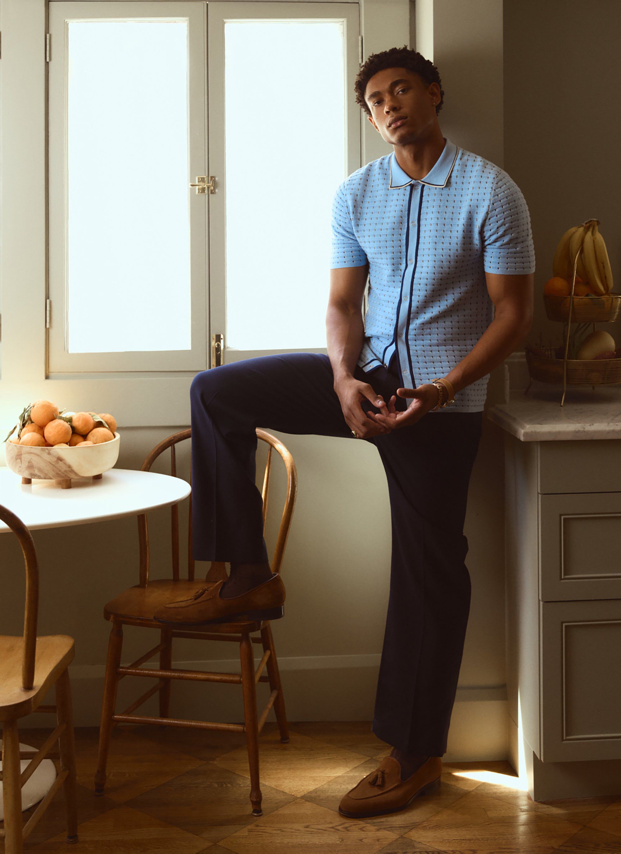 A model in a sunlit kitchen wears the blue Broken Stripe Knitted Shirt from Percival with navy trousers and brown loafers.