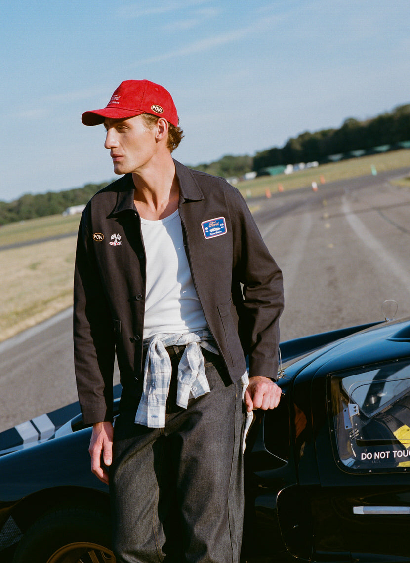 Navy GT40 embroidered canvas jacket and red trucker cap from the Ford x Percival collection on a racing track.