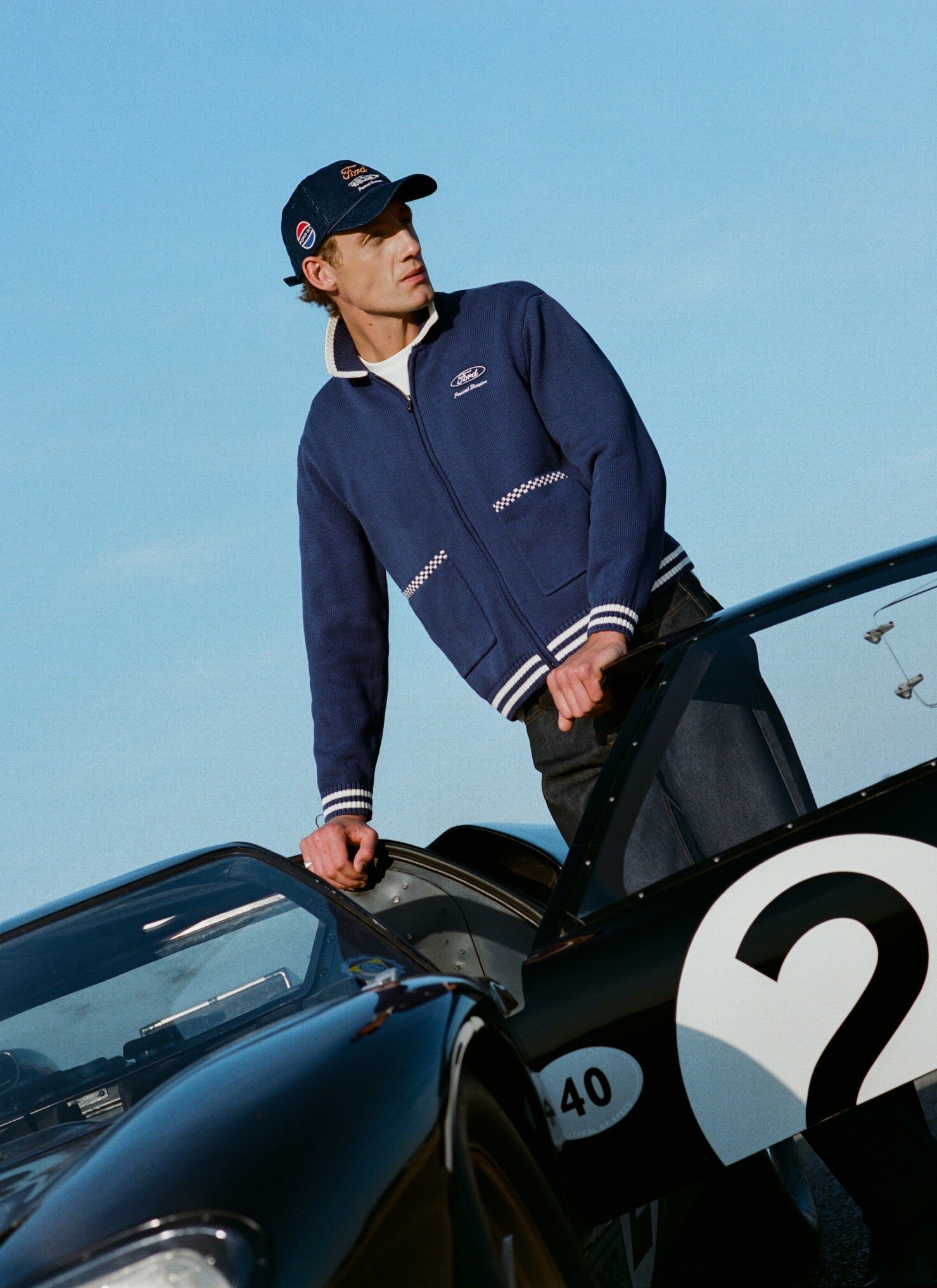 Navy Ford x Percival Gardner Knitted Cardigan styled with dark denim jeans and a cap by a vintage racing car.