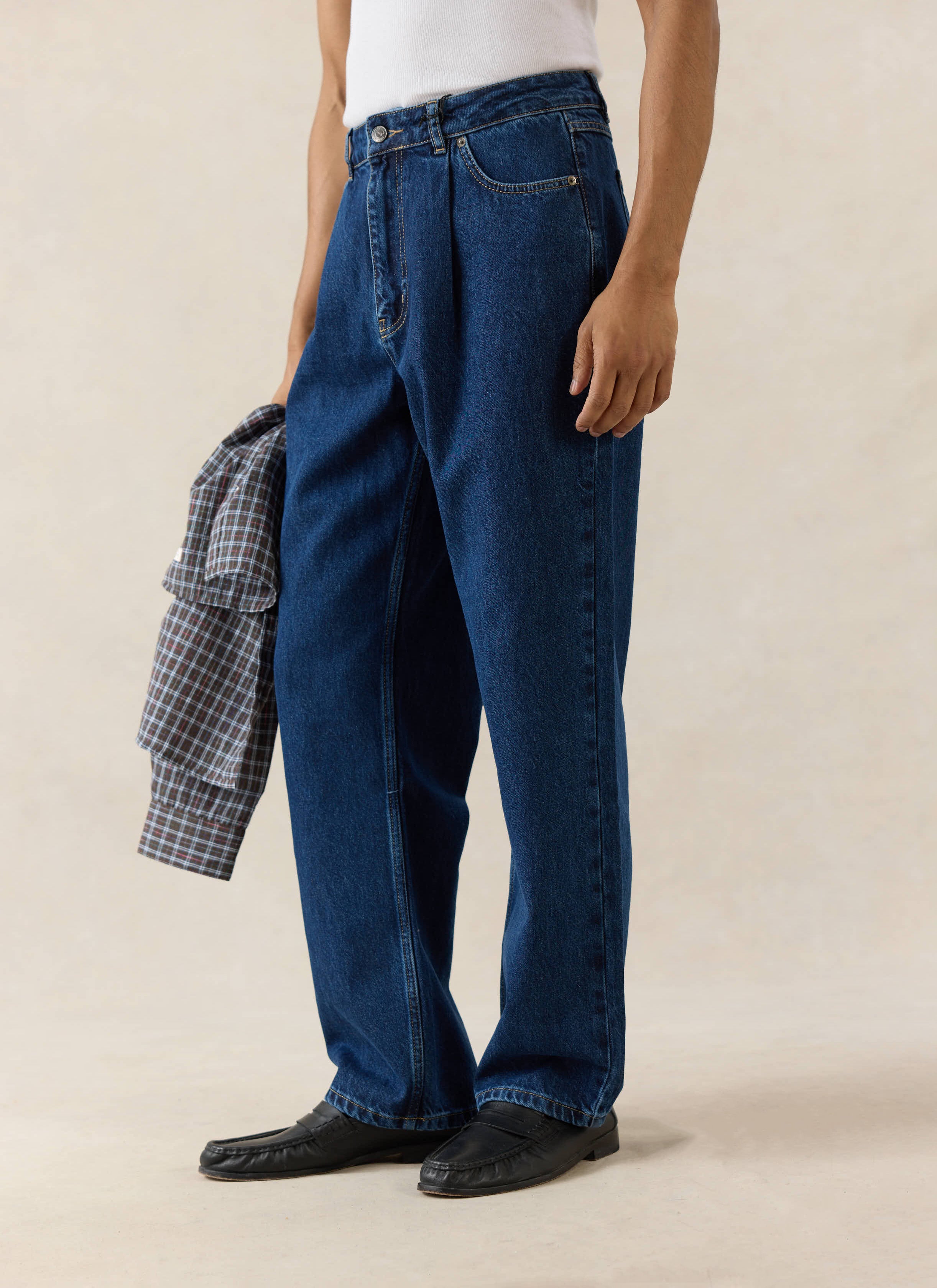 Percival Pleated Denim Jeans in 12oz mid-wash blue cotton, styled with a white tank top, black loafers, and a check shirt.