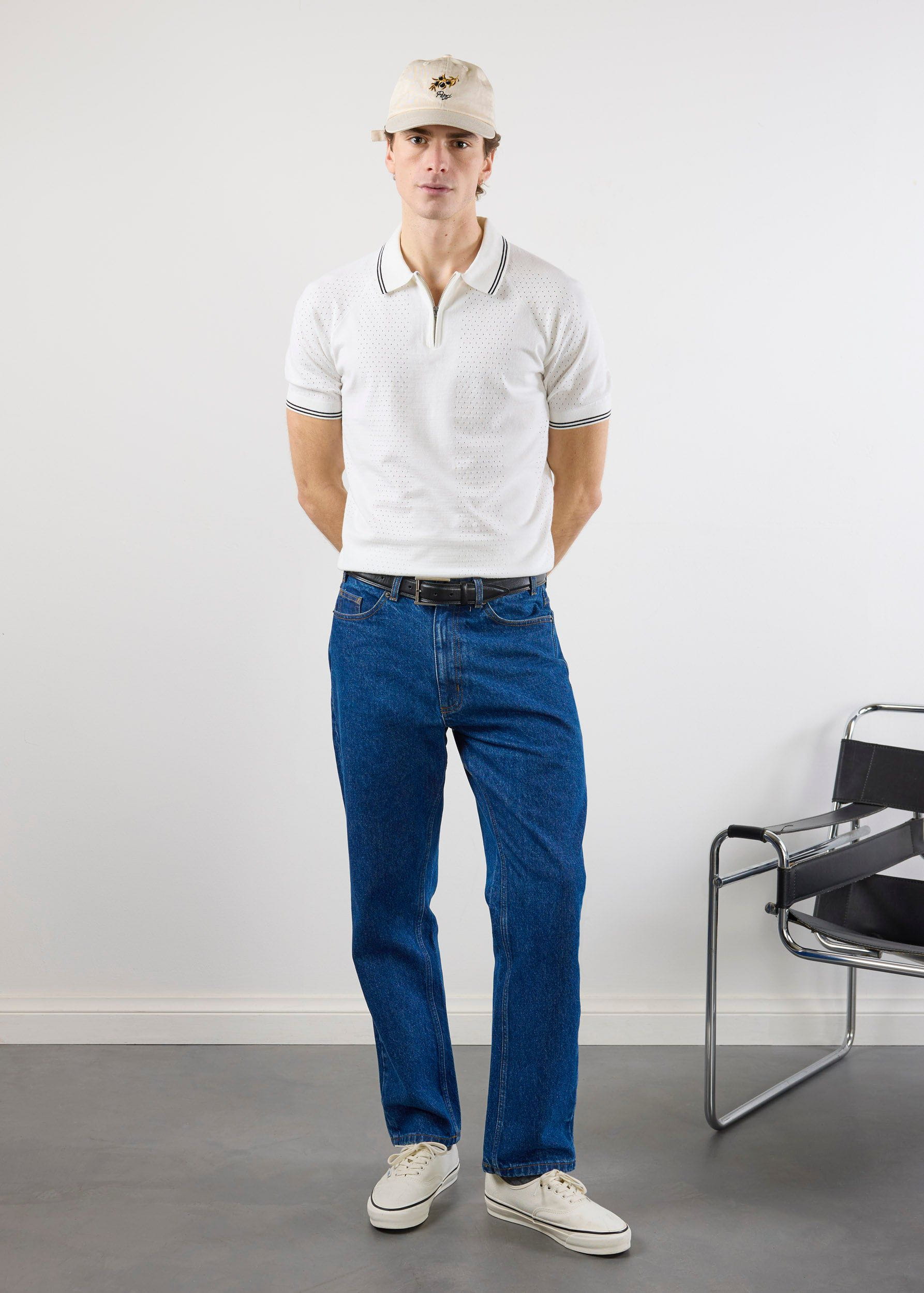 A model wears the Percival Rossi Knitted Polo in white, styled with dark blue jeans, a cap, and white trainers.
