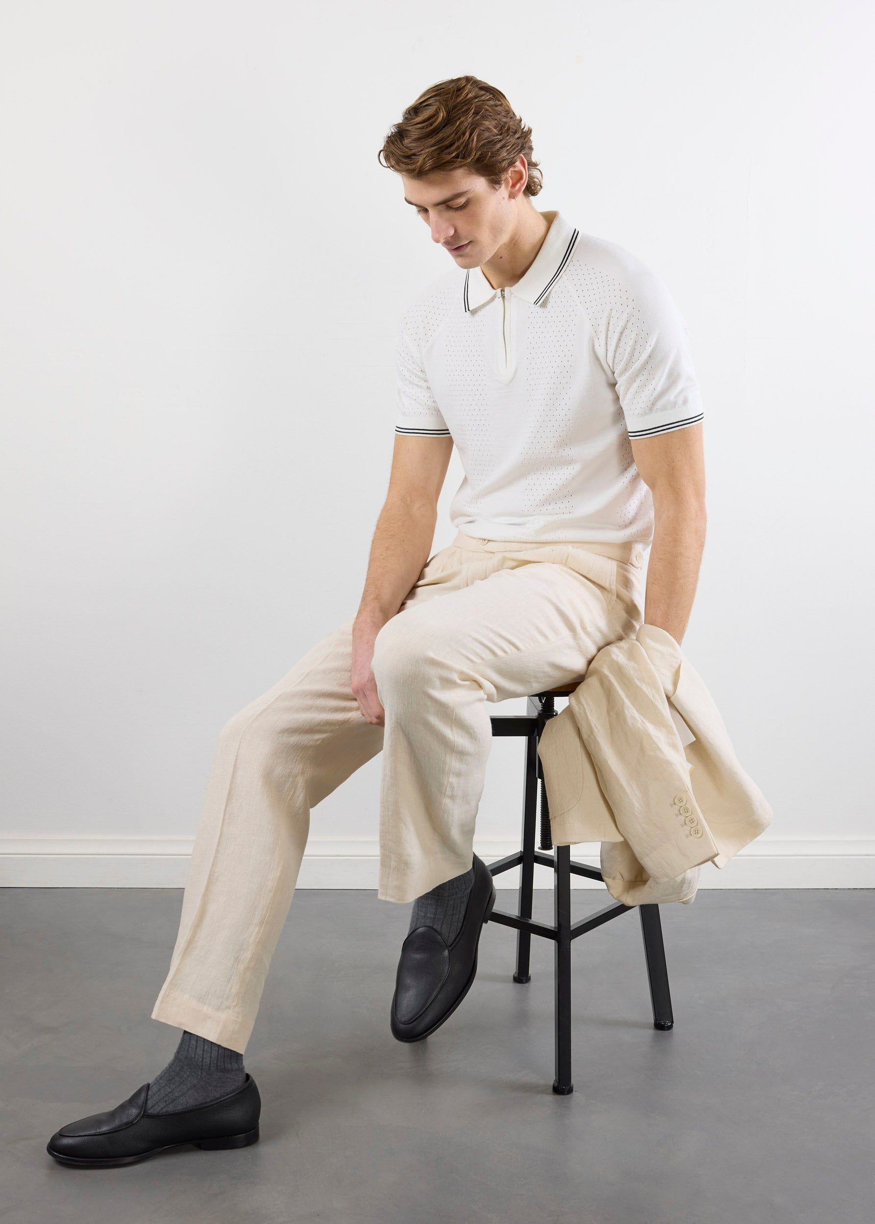 A model sits on a stool wearing the Percival Rossi Knitted Polo in white, paired with ecru trousers and a matching jacket.