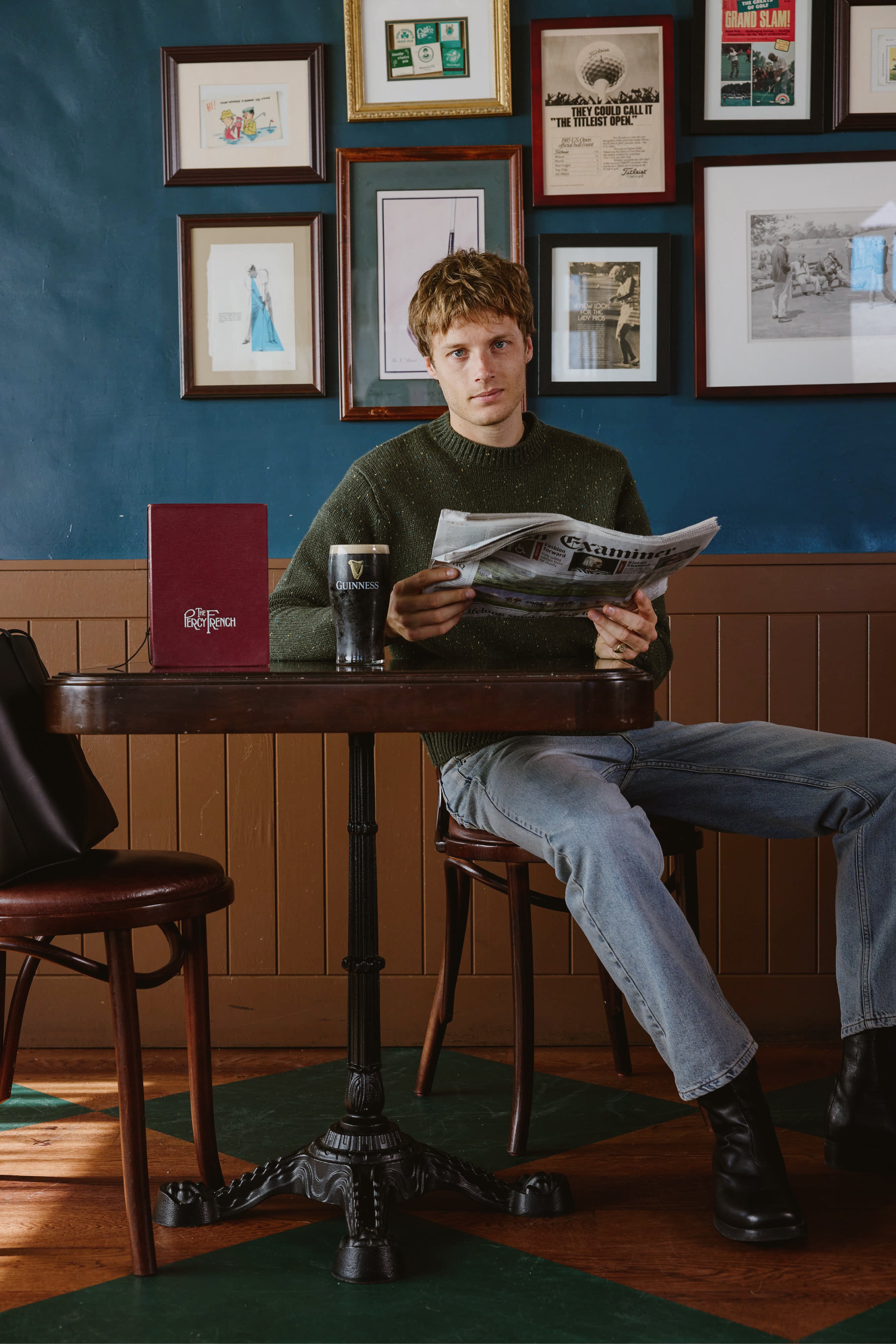A model wears the Percival forest green Rhodes jumper, sitting at a pub table reading a newspaper with a pint of Guinness.