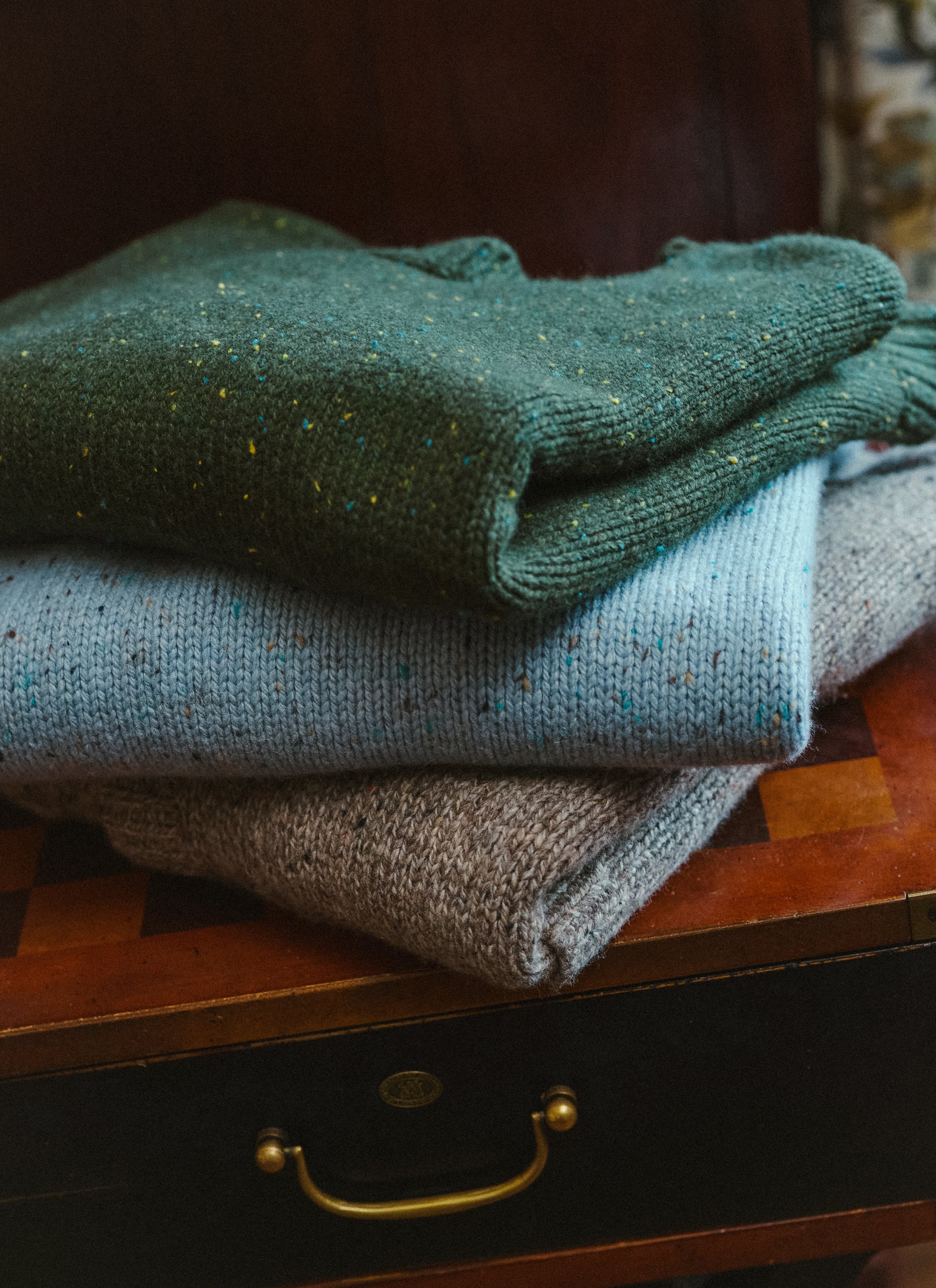 A folded stack of Percival knitwear featuring the Rhodes crew neck jumpers and cardigan in forest green, blue, and oak.