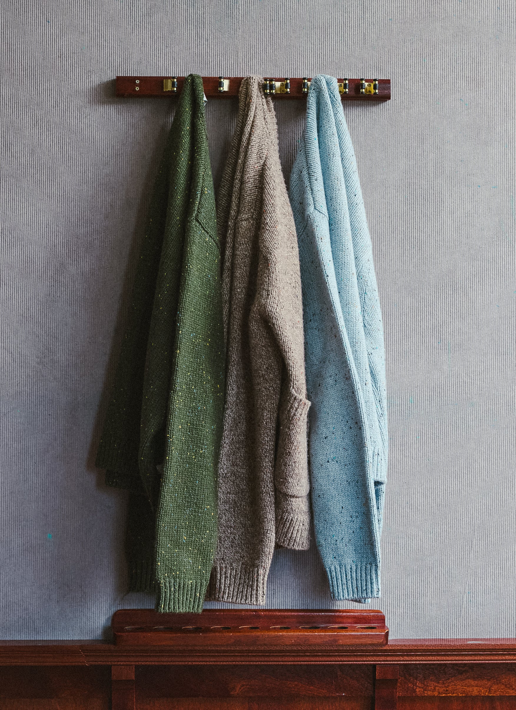 Three Percival knitwear pieces in forest green, oak, and blue hanging on a wooden peg rail.