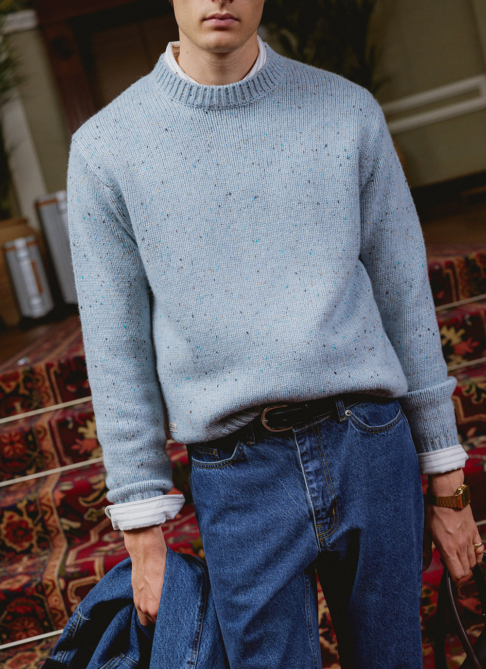 A model wears the blue flecked Rhodes crew neck jumper from Percival over a white shirt, paired with blue jeans.