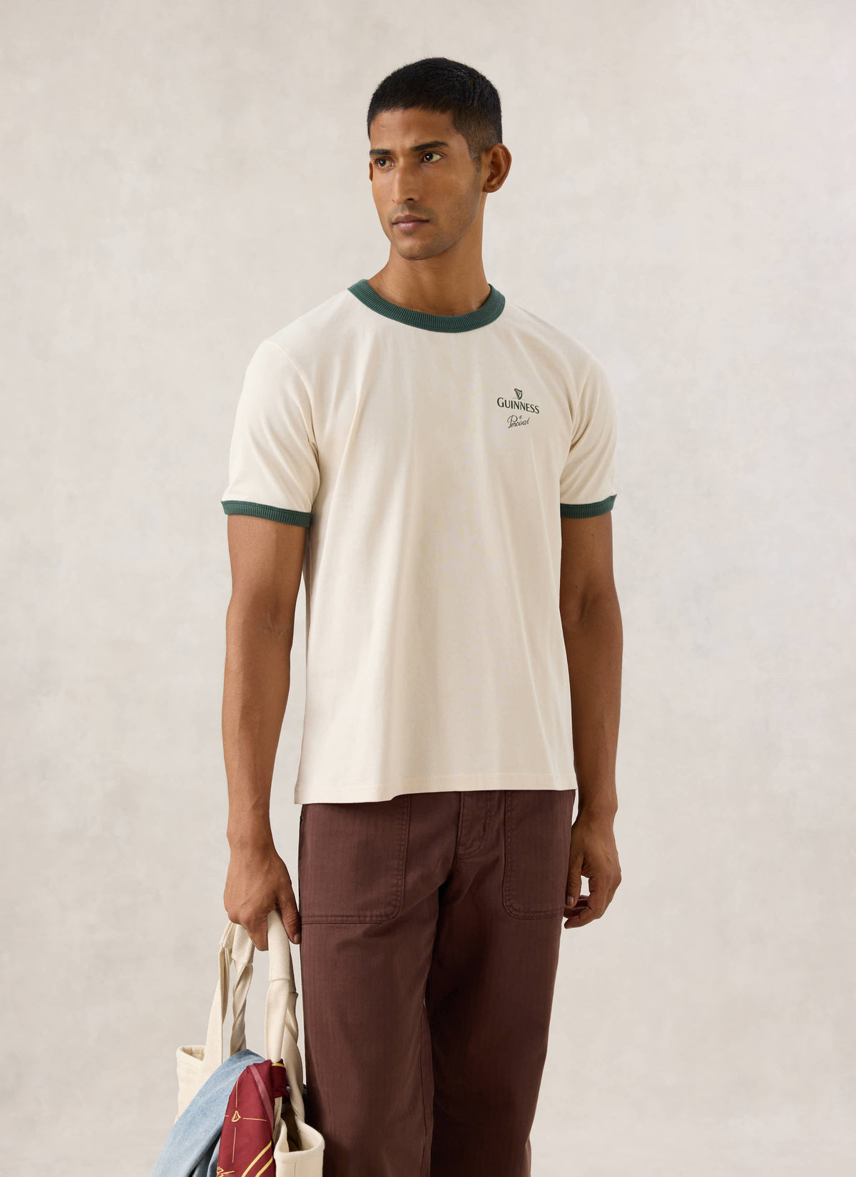 Model wearing the Guinness x Percival cream ringer T-shirt with forest green trim, brown trousers, and holding a tote bag.