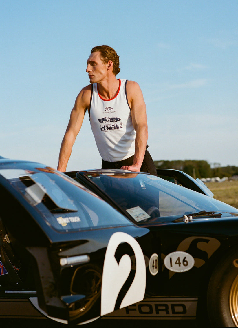 Man wears a white Ford x Percival ringer vest with red trim, standing beside a vintage black Ford racing car outdoors.